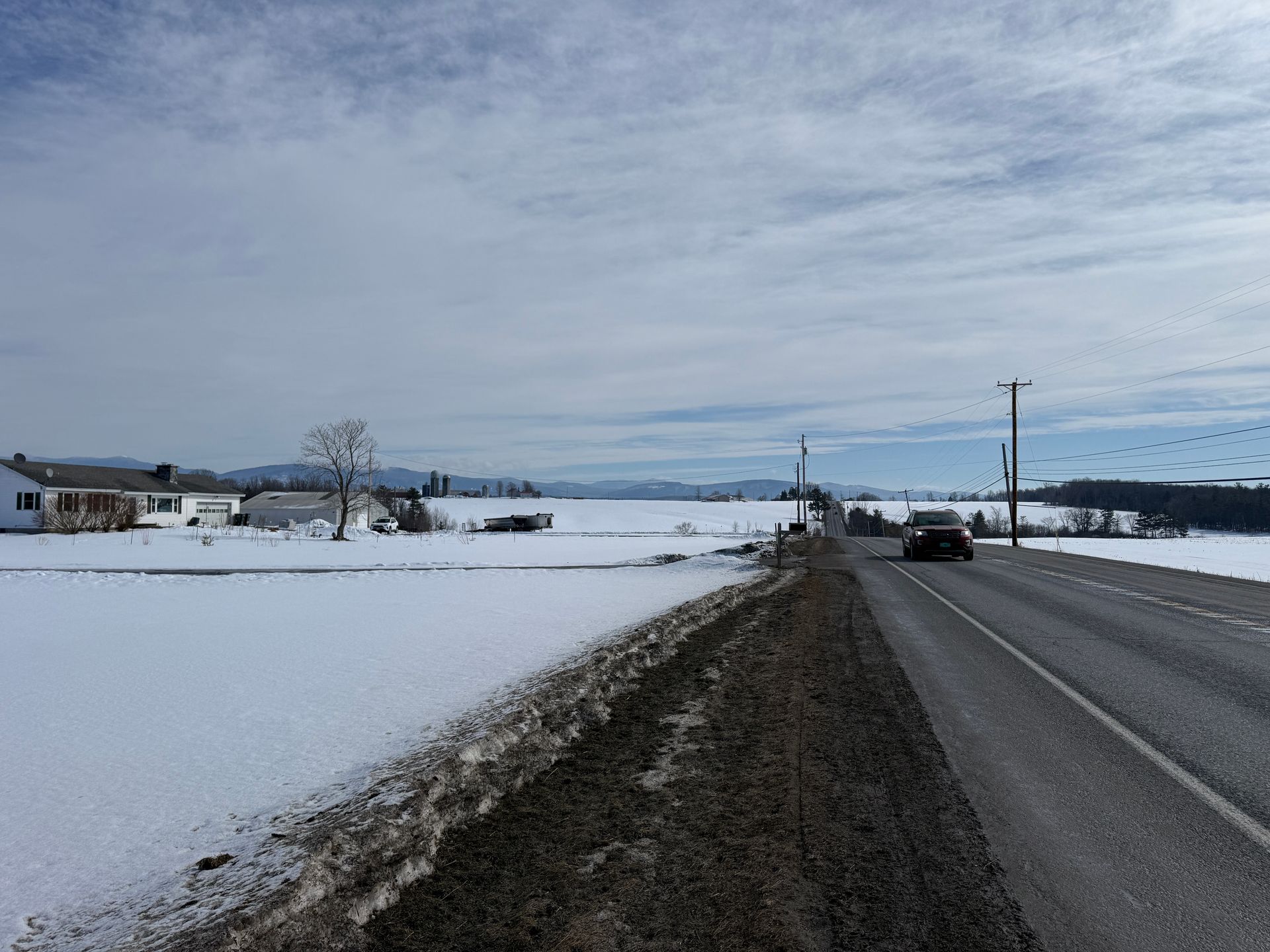 Snowy landscape with a road and a car driving, houses in the distance, and a cloudy sky.