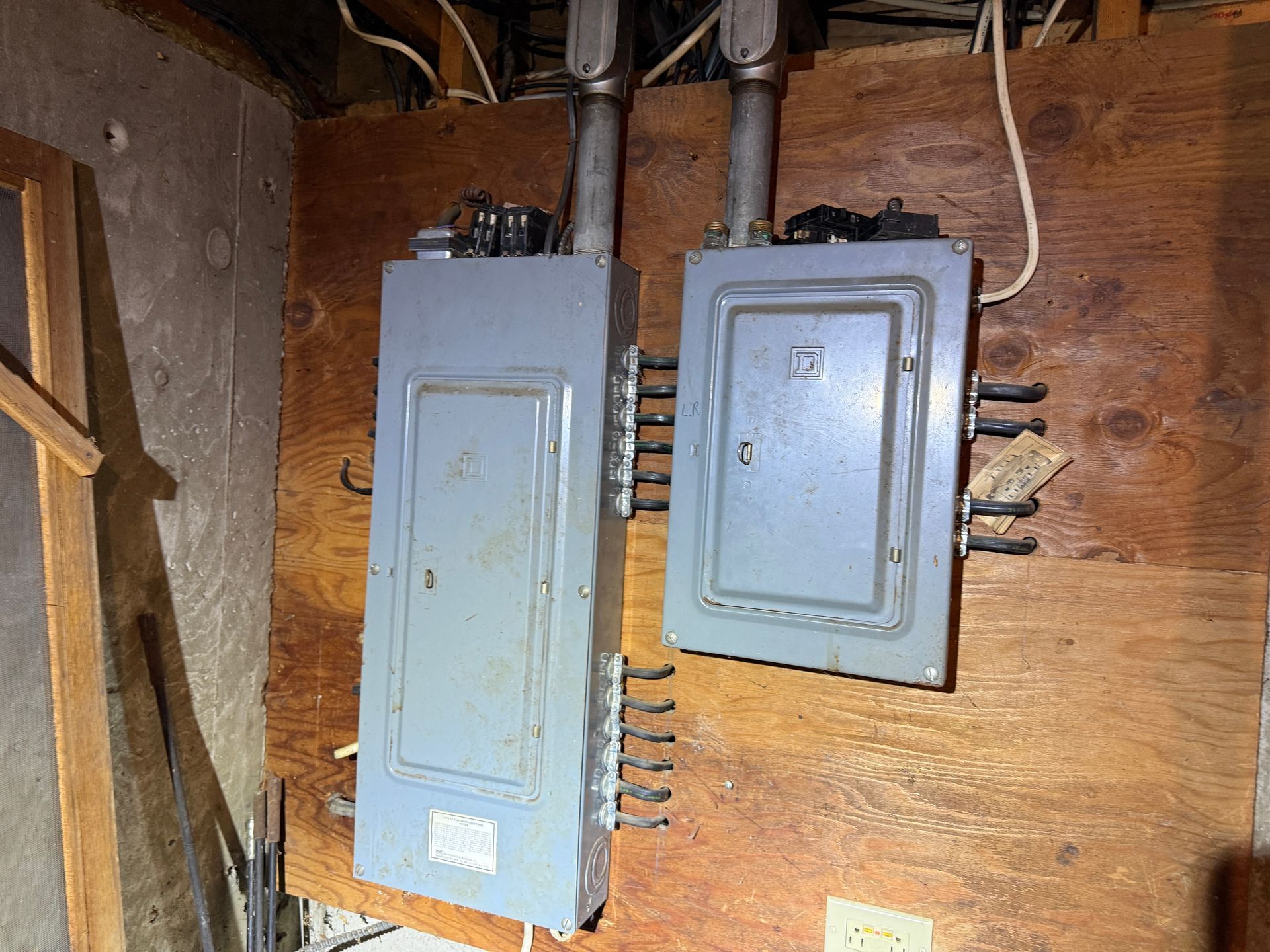 Two gray electrical breaker boxes mounted on a wood-paneled wall with conduit pipes.