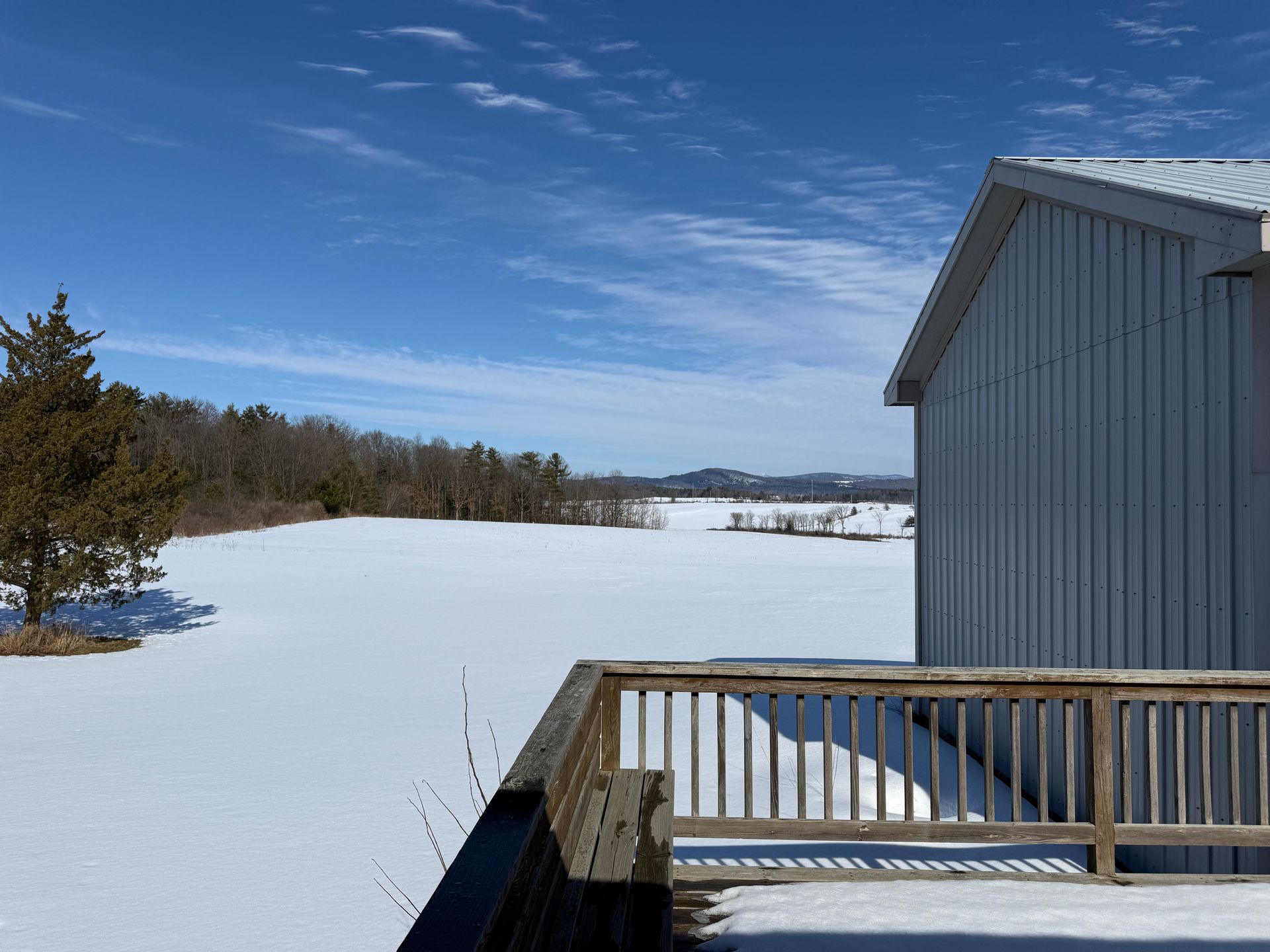 Snowy field and trees under a blue sky, viewed from a wooden deck next to a gray building.