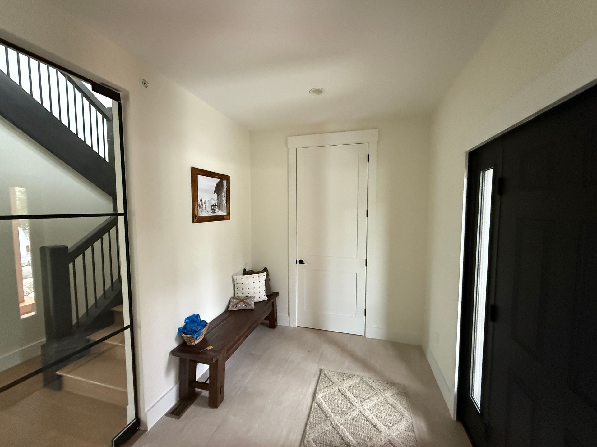 A bright entryway with a dark door, a wooden bench against a wall, a staircase with a black railing, and a patterned rug.
