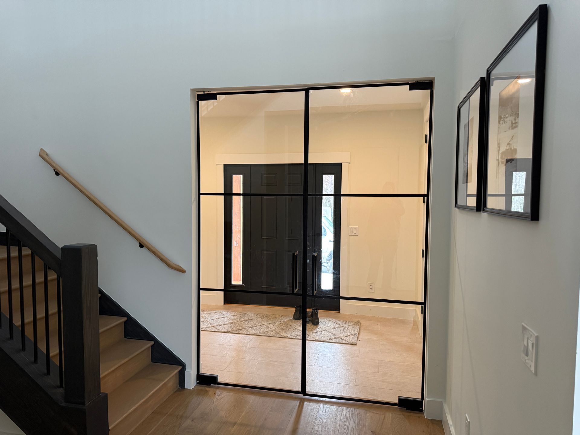 A hallway with a wooden staircase and a black-framed glass double door leading to a black exterior entryway.