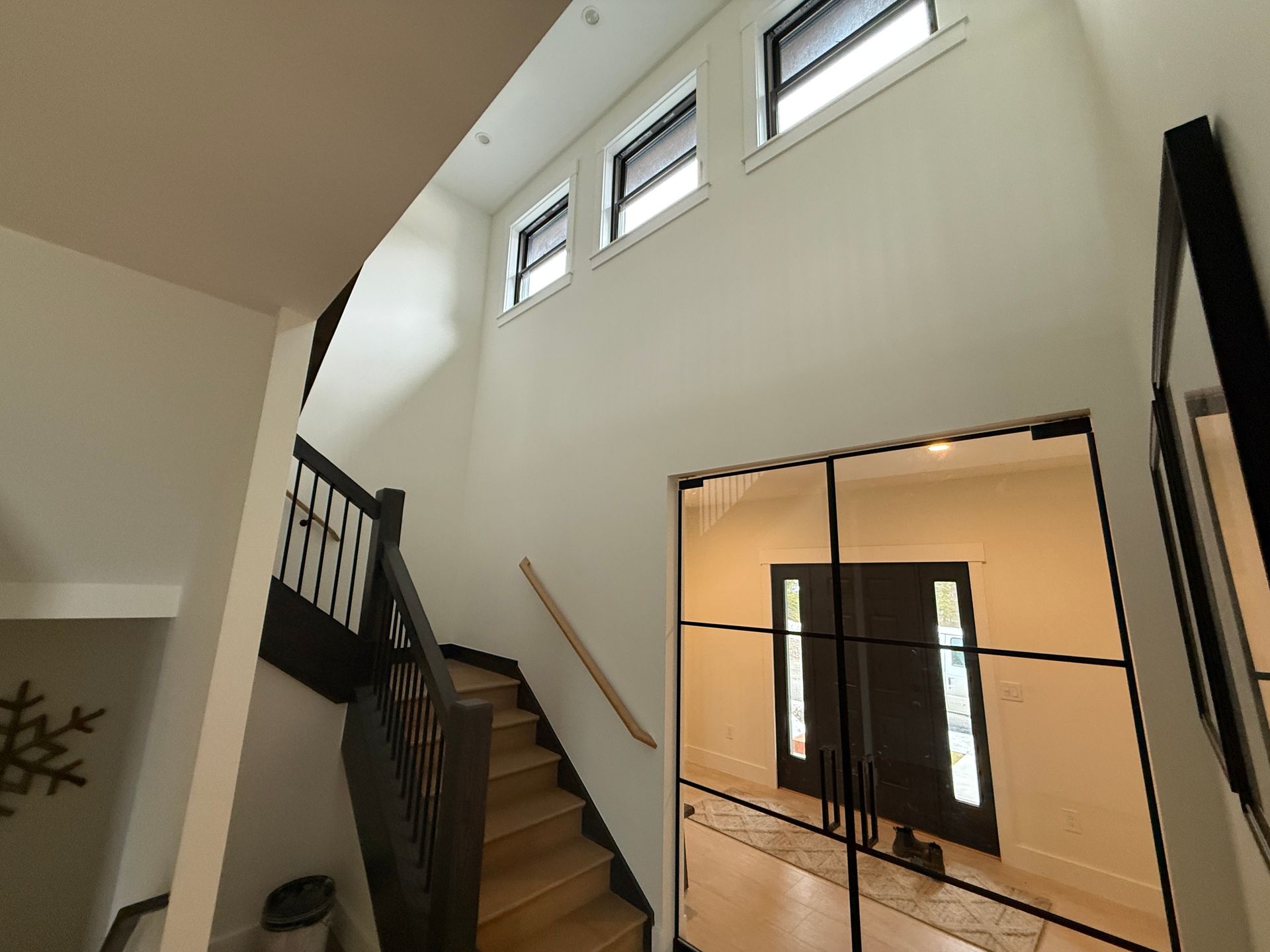 A bright indoor entryway with light walls, a wooden staircase with black railings, and a black-framed glass door.