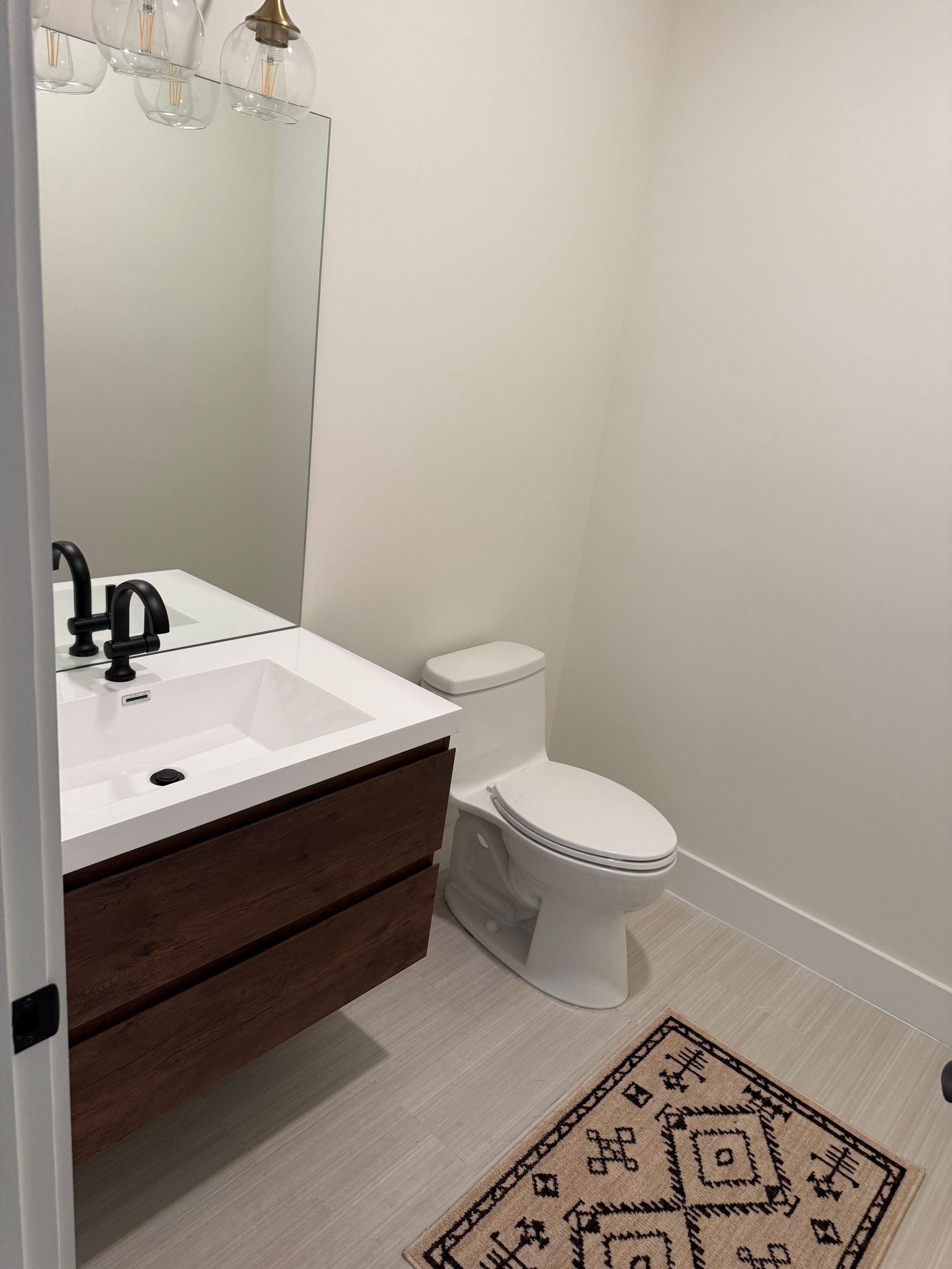 A modern half-bathroom with a floating wood vanity, white sink, matte black faucet, wall mirror, and patterned floor rug.