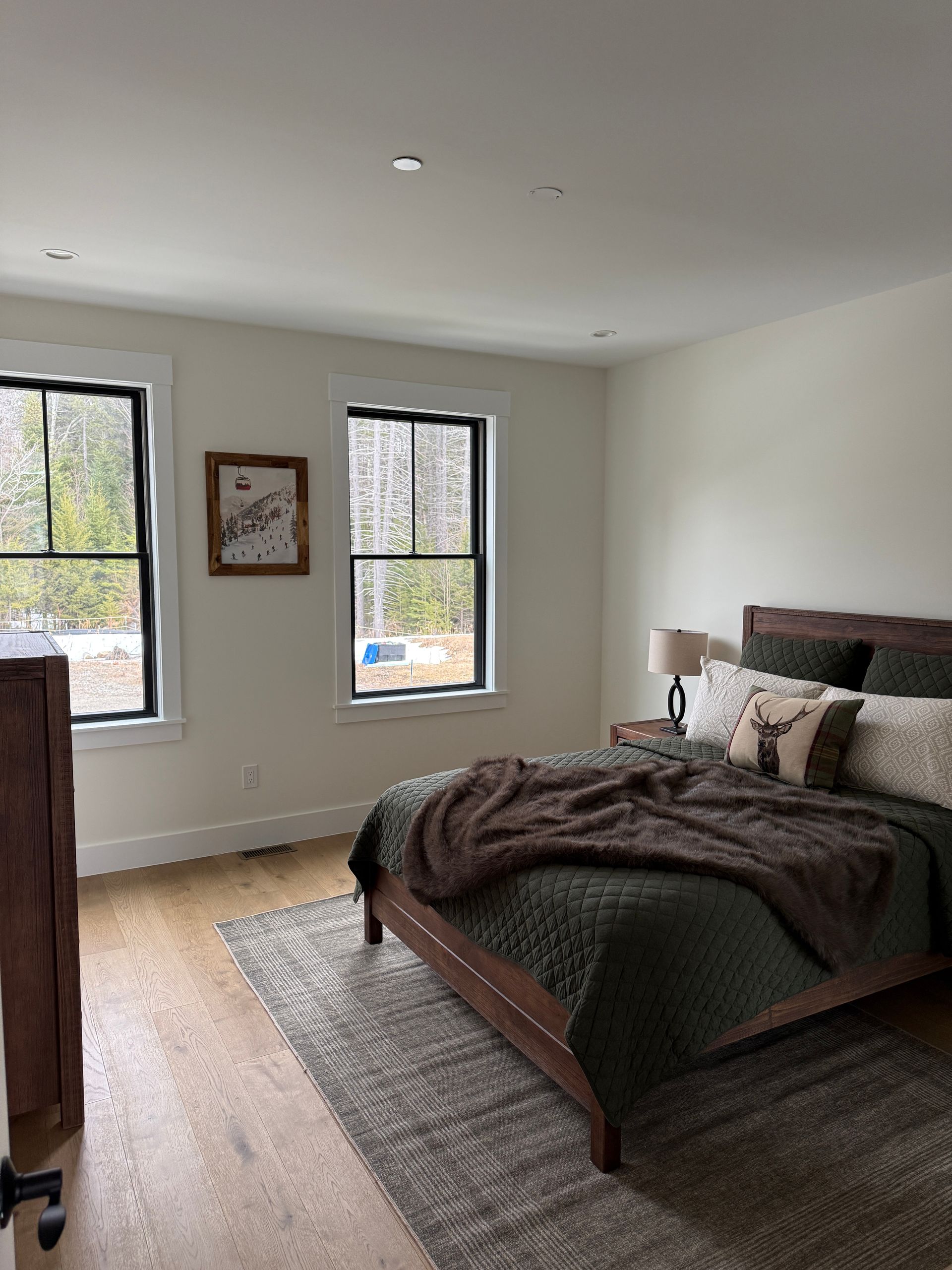 A neutral-toned bedroom with a wooden bed, dark green bedding, a patterned rug, and two windows facing an outdoor view.