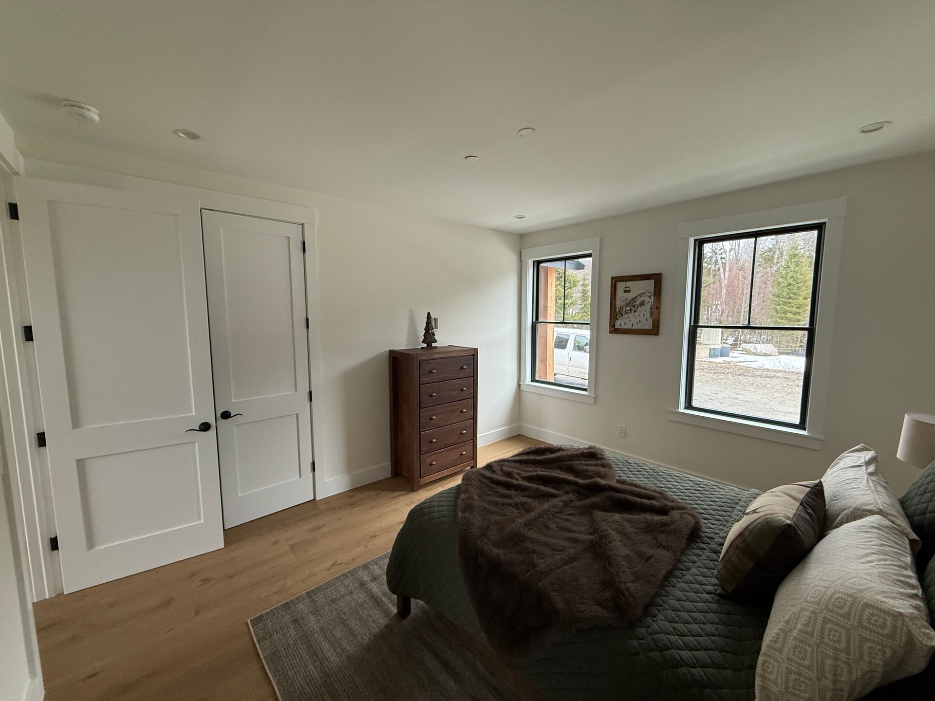 A modern bedroom with white walls, light wood floors, a bed with a dark quilt, two windows, and closed white double doors.