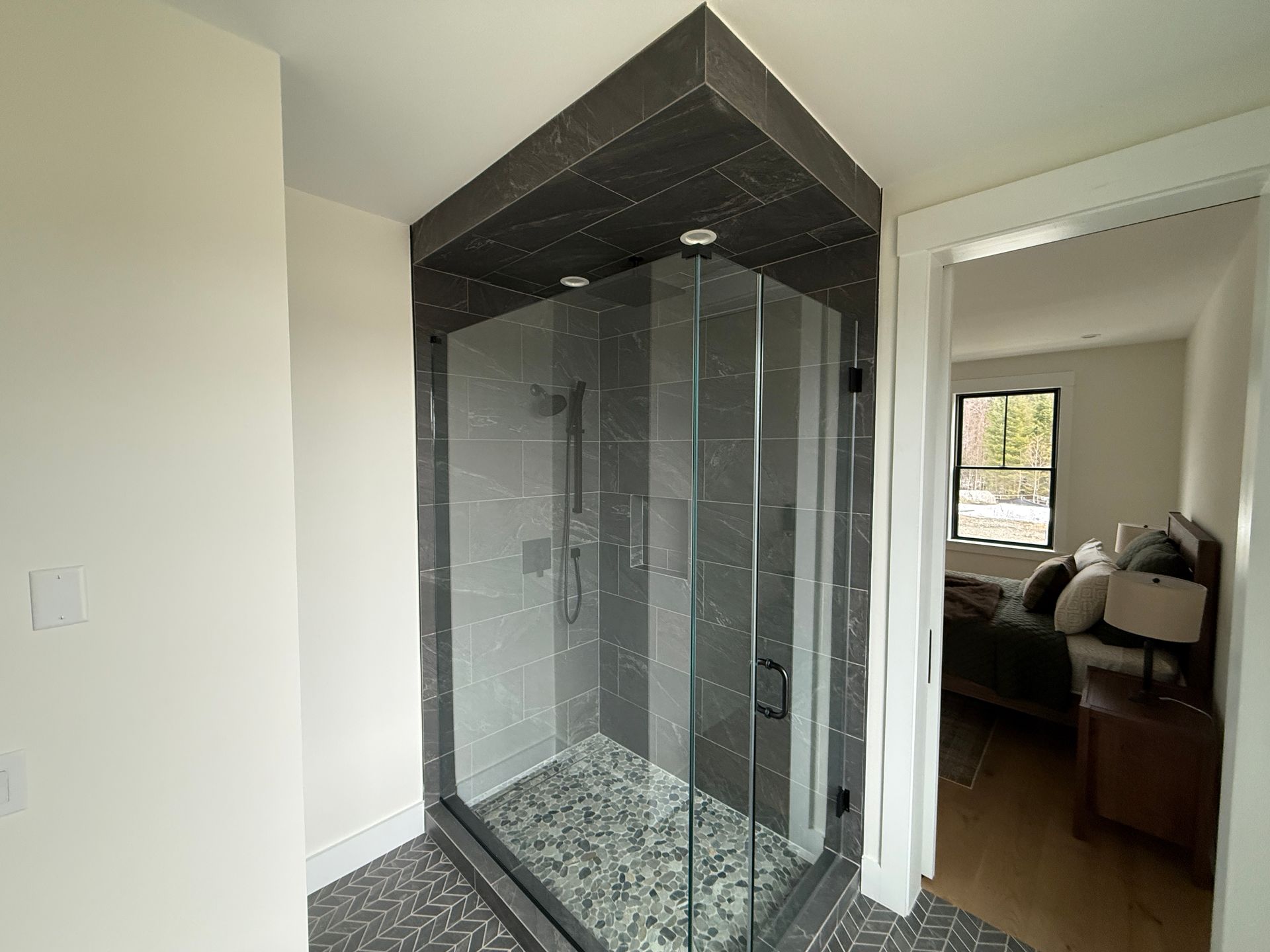A glass shower enclosure with dark gray tile walls and a pebble floor, adjacent to a doorway leading to a bedroom.