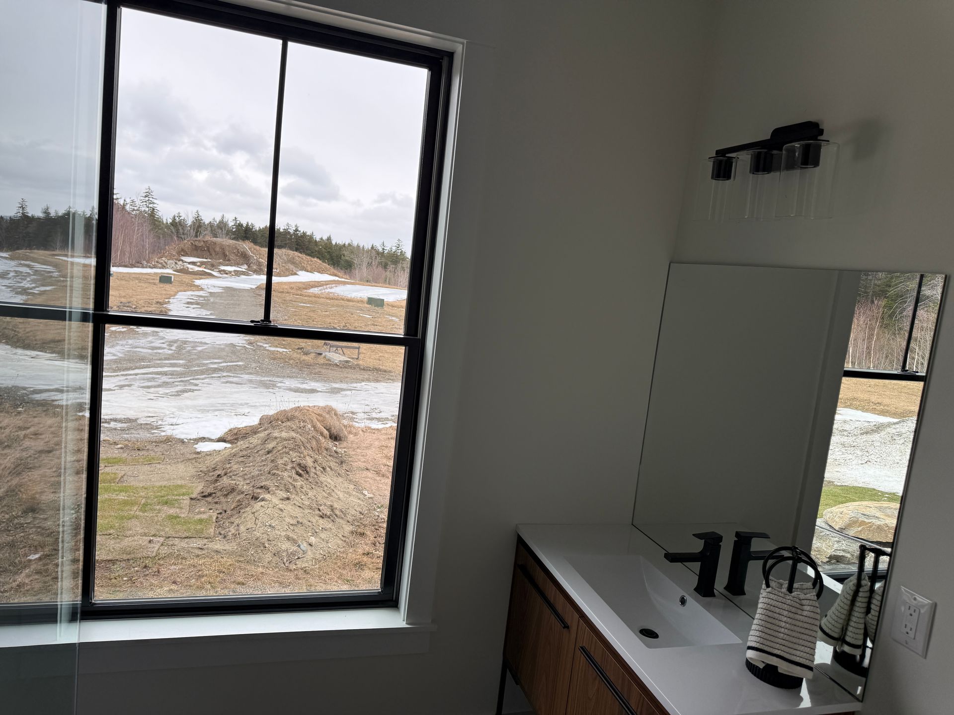 A bathroom vanity with a white countertop and black fixtures next to a window overlooking a snowy, rural landscape.