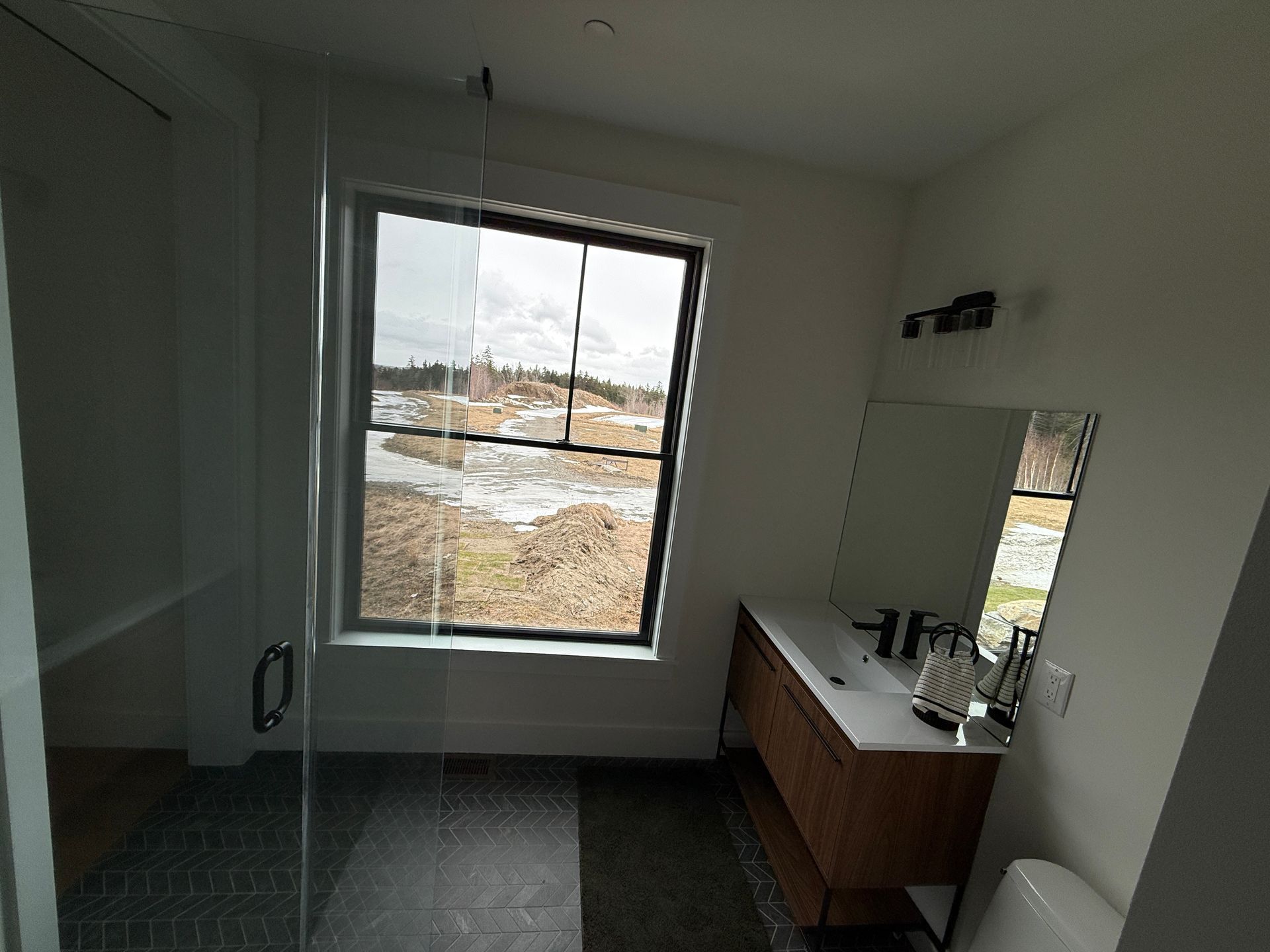 A modern bathroom with a glass-enclosed shower, wood vanity, mirror, and a window overlooking a landscape.