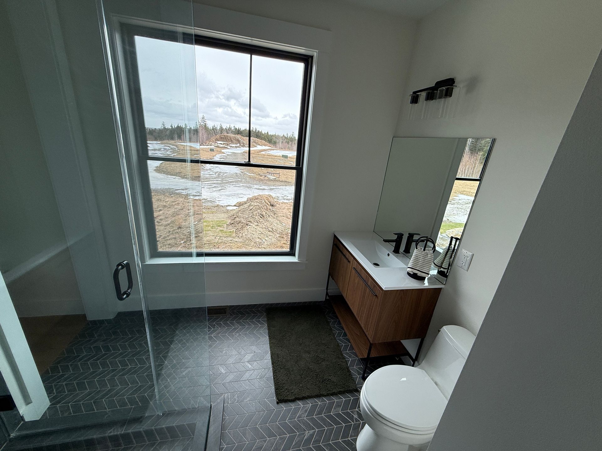A modern bathroom with a glass shower, a vanity, a toilet, and a large window looking out onto a landscape.