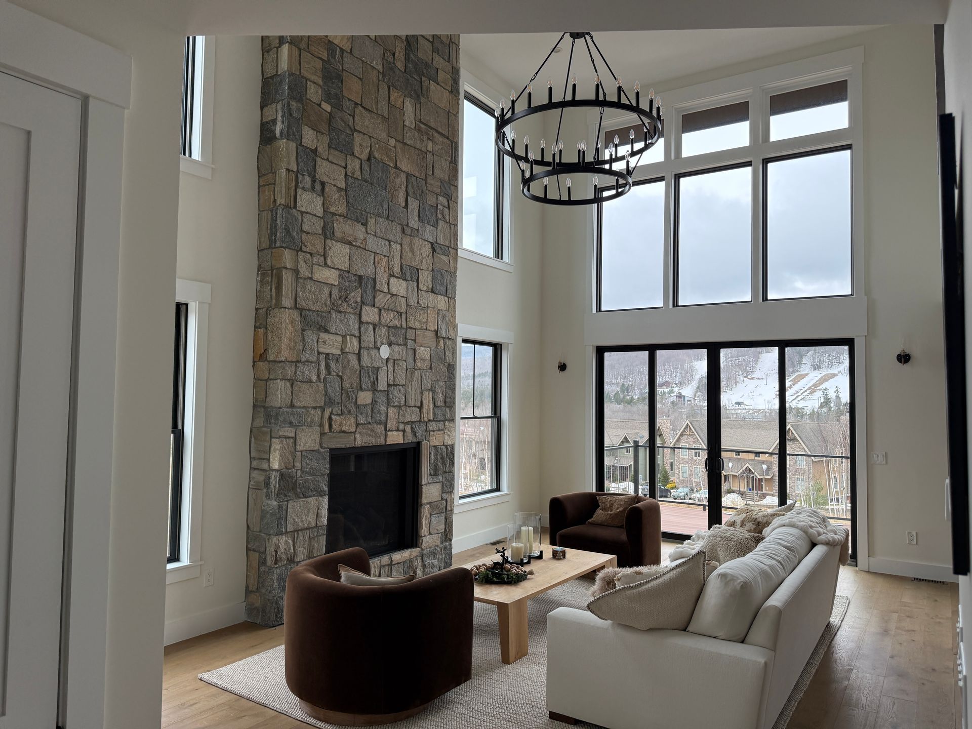 A modern living room with high ceilings, stone fireplace, white sofa, two brown chairs, and large windows with mountain view.