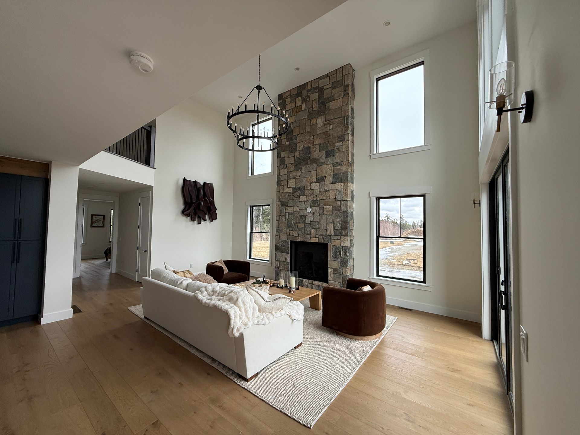 A bright, modern living room with a stone fireplace, a white sofa, two brown chairs, and a chandelier.