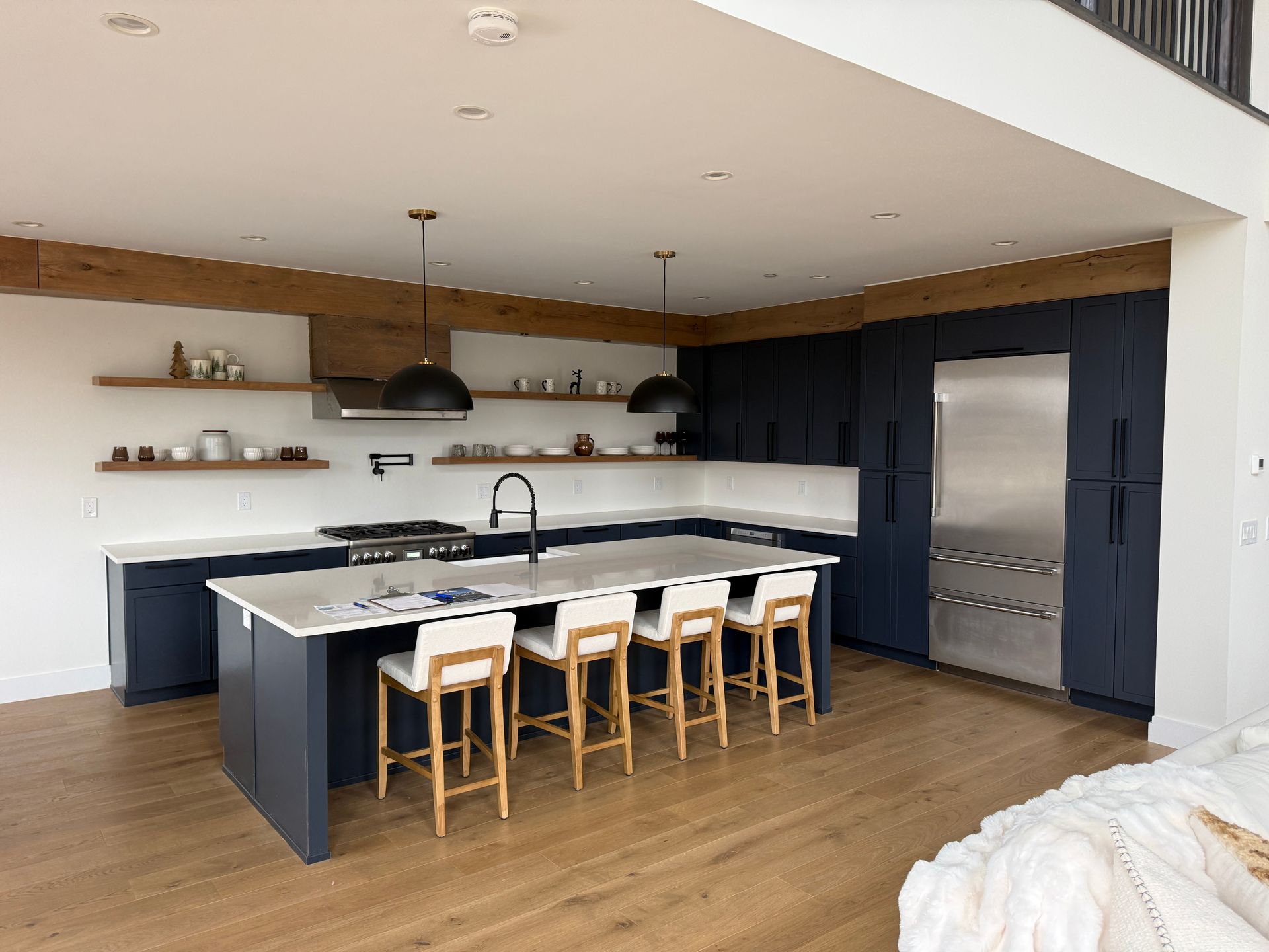 Modern kitchen with dark navy cabinets, wood floating shelves, a large white-topped island, and four wooden bar stools.