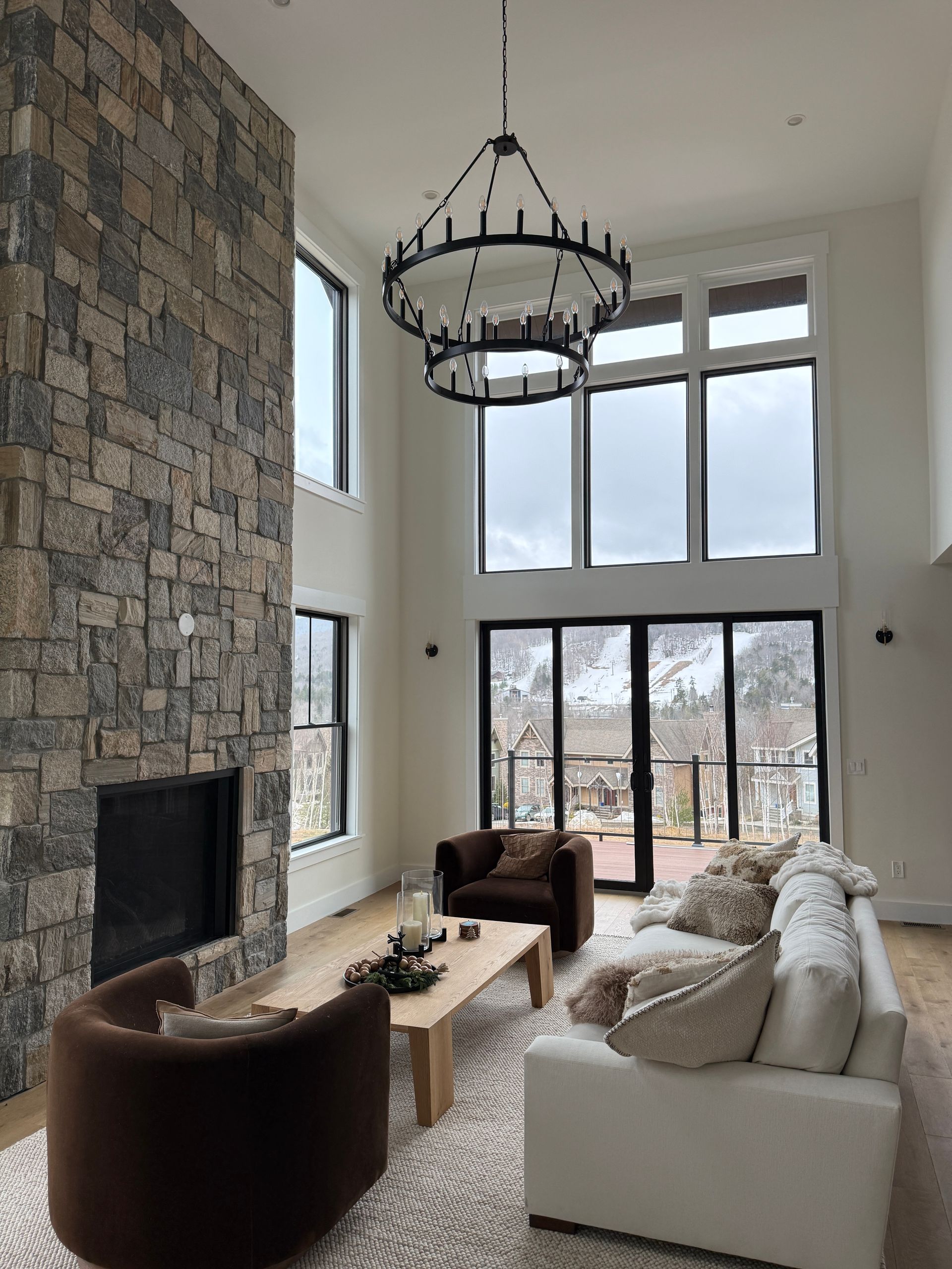 Living room with high ceilings, large stone fireplace, chandelier, white sofa, and two armchairs facing a snowy mountain.
