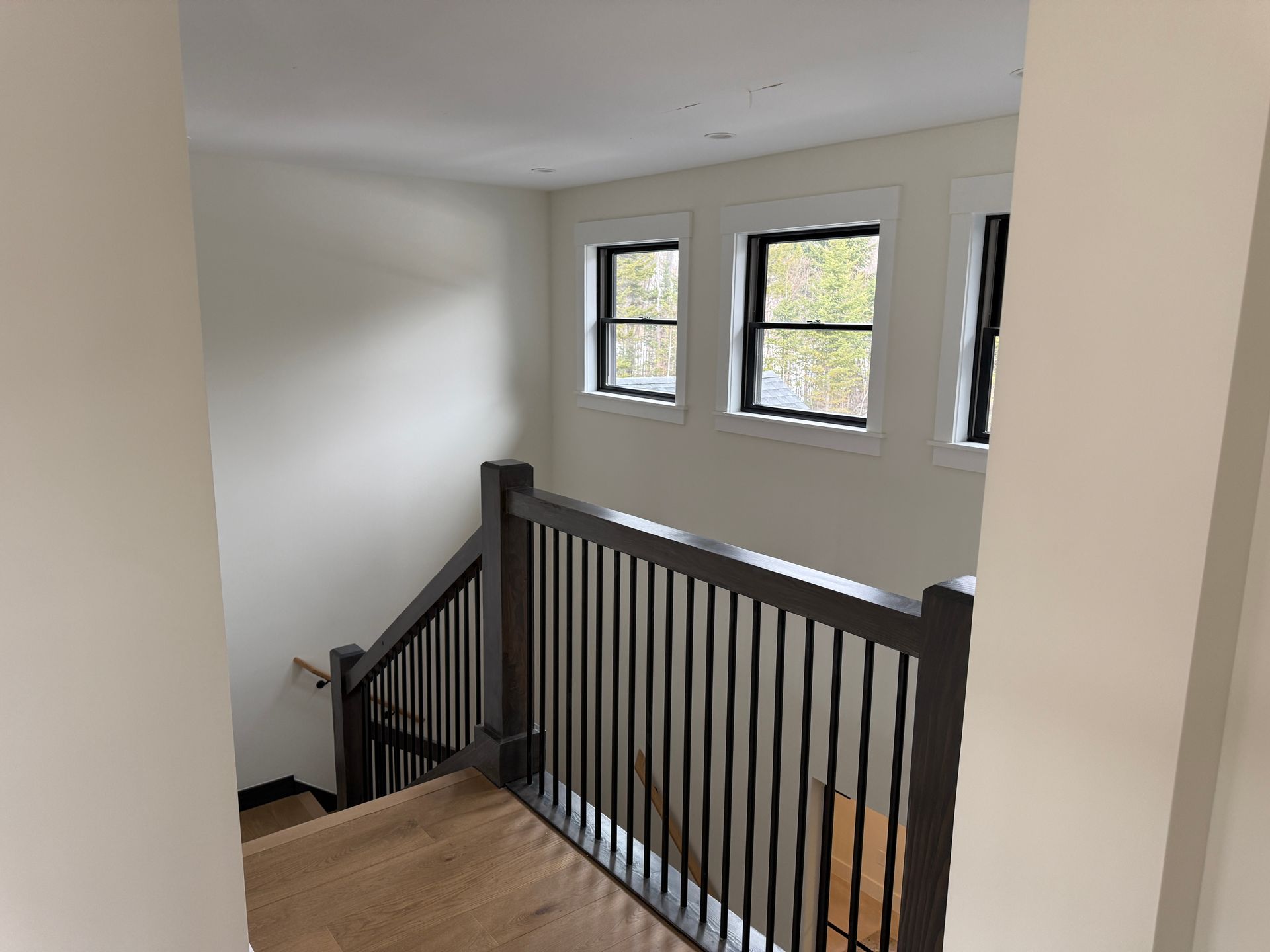 A wooden staircase with a dark railing and white walls, viewed from the top landing with three windows overlooking trees.