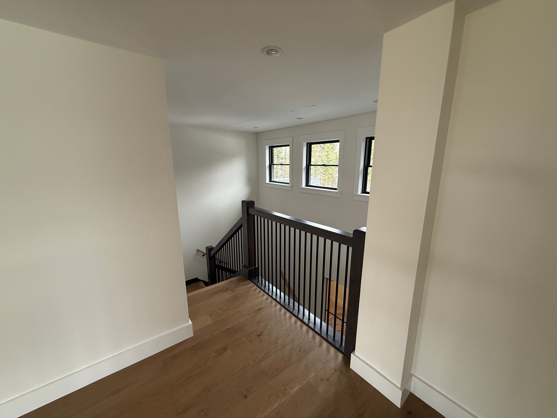 A landing with light wood floors and white walls, featuring a dark wooden staircase railing and three bright windows.