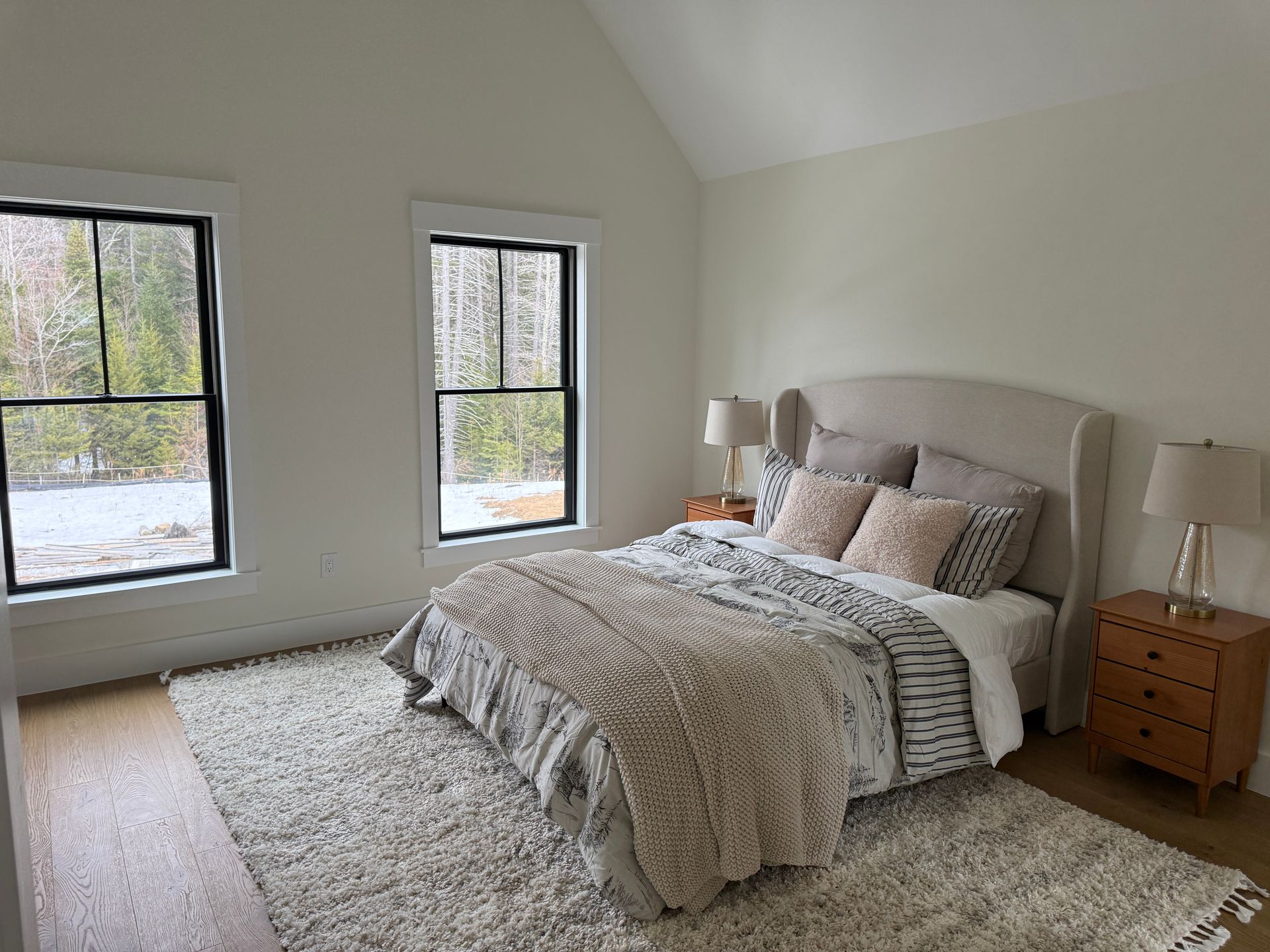 A bedroom with a neutral-colored bed, a textured rug, two wooden nightstands, and two windows overlooking a snowy forest.