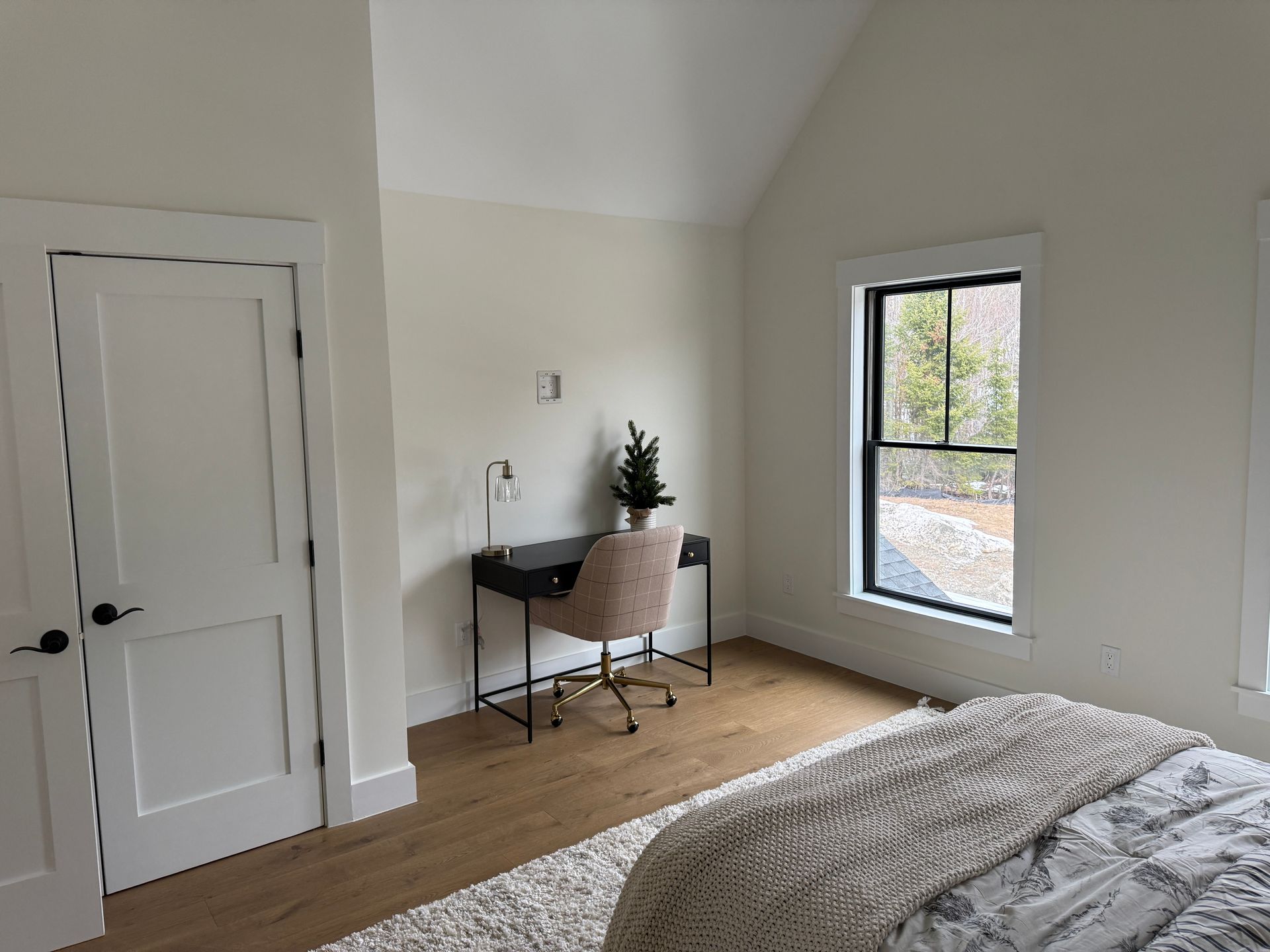 A bedroom with white walls, wood floors, a desk with a chair under a window, and a bed with a textured blanket.
