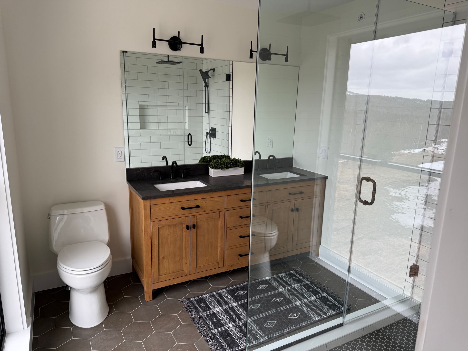 A bathroom with a wooden vanity, a white toilet, gray hexagonal floor tiles, and a walk-in glass shower.