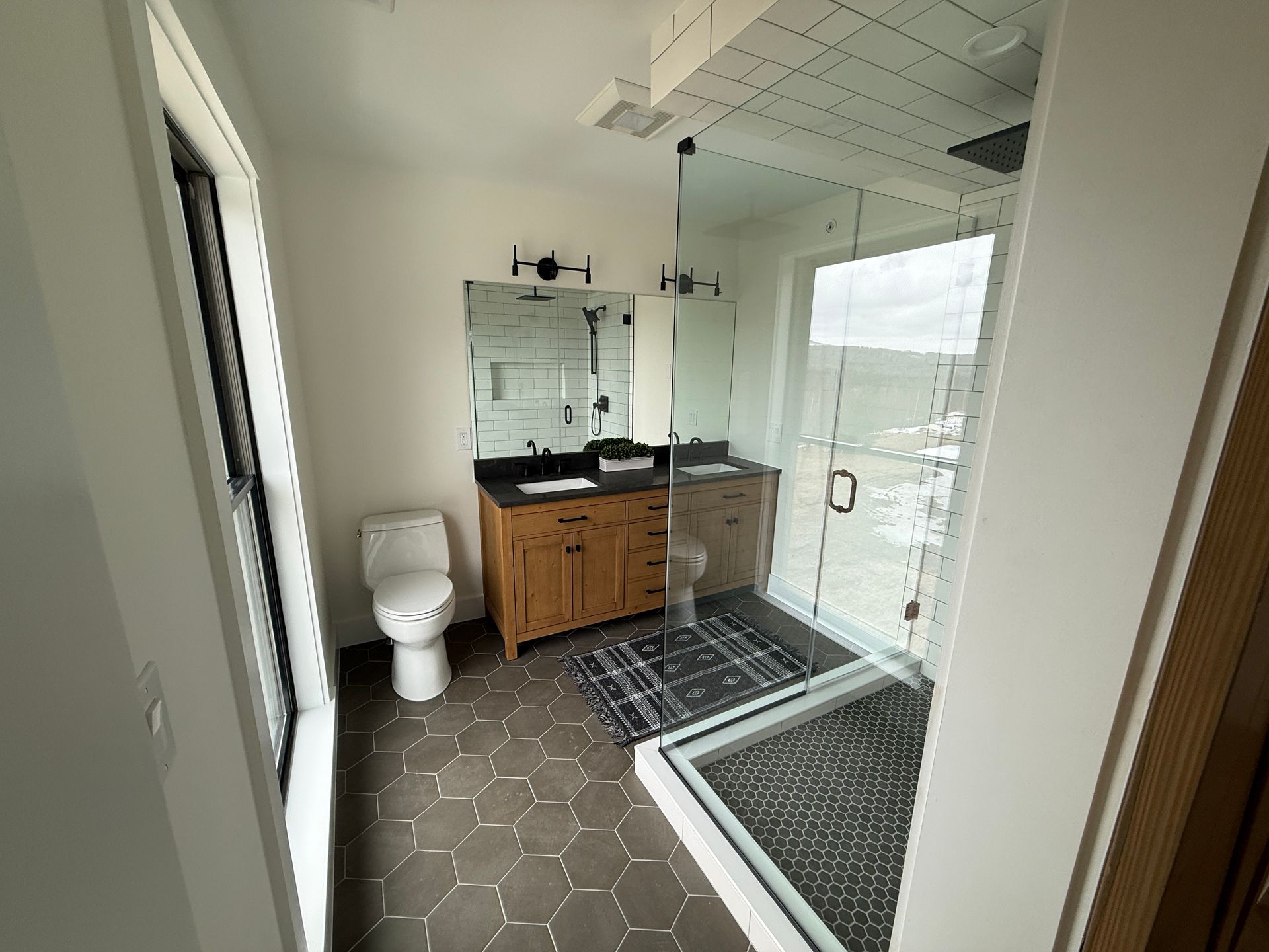 A modern bathroom with dark hexagonal floor tiles, a wood vanity, white toilet, and a glass-enclosed walk-in shower.