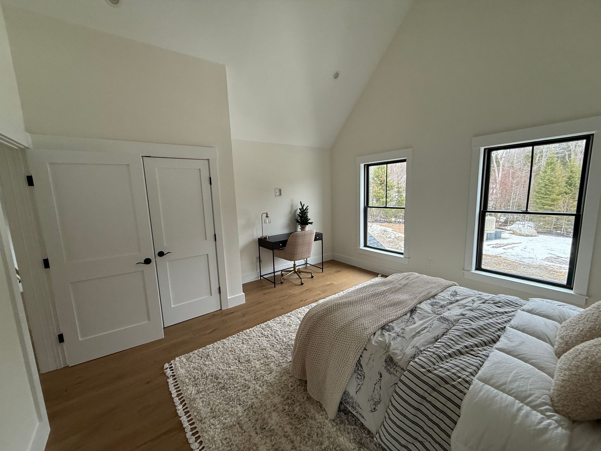 A bright bedroom with a white vaulted ceiling, a bed with a textured rug, two windows, and a small desk in the corner.