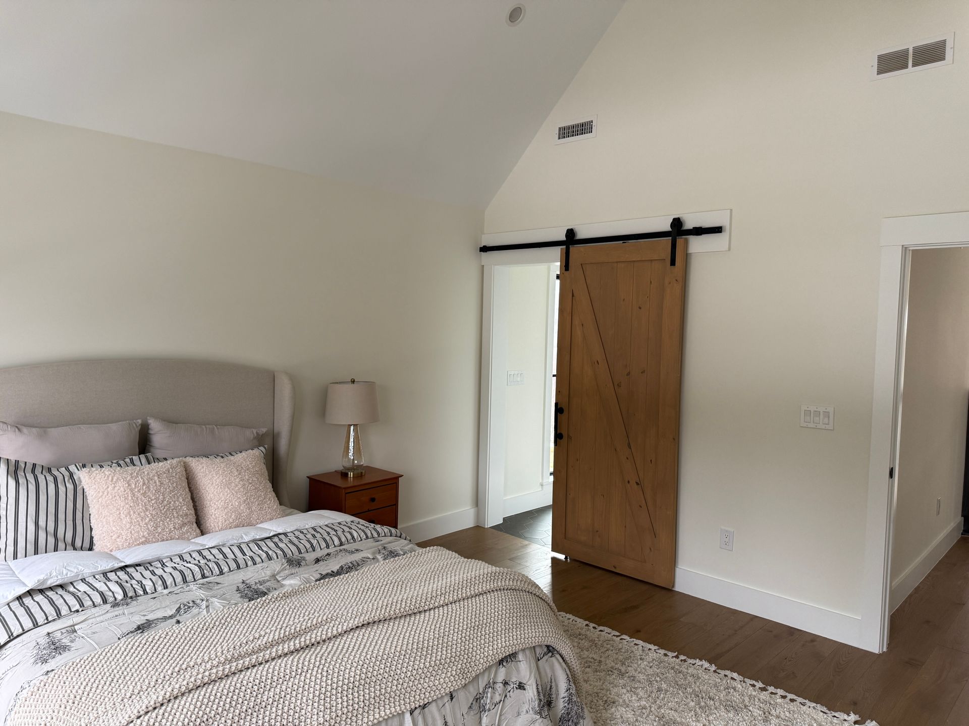 A bedroom with a beige upholstered bed, patterned bedding, wooden nightstand, and a light wood barn door in a white room.