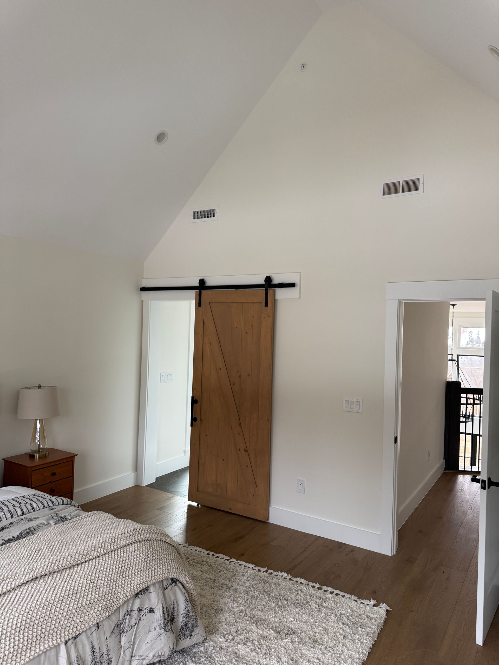 A bedroom with a vaulted ceiling, a wooden barn door, a bedside lamp, and an open doorway leading to a hallway.