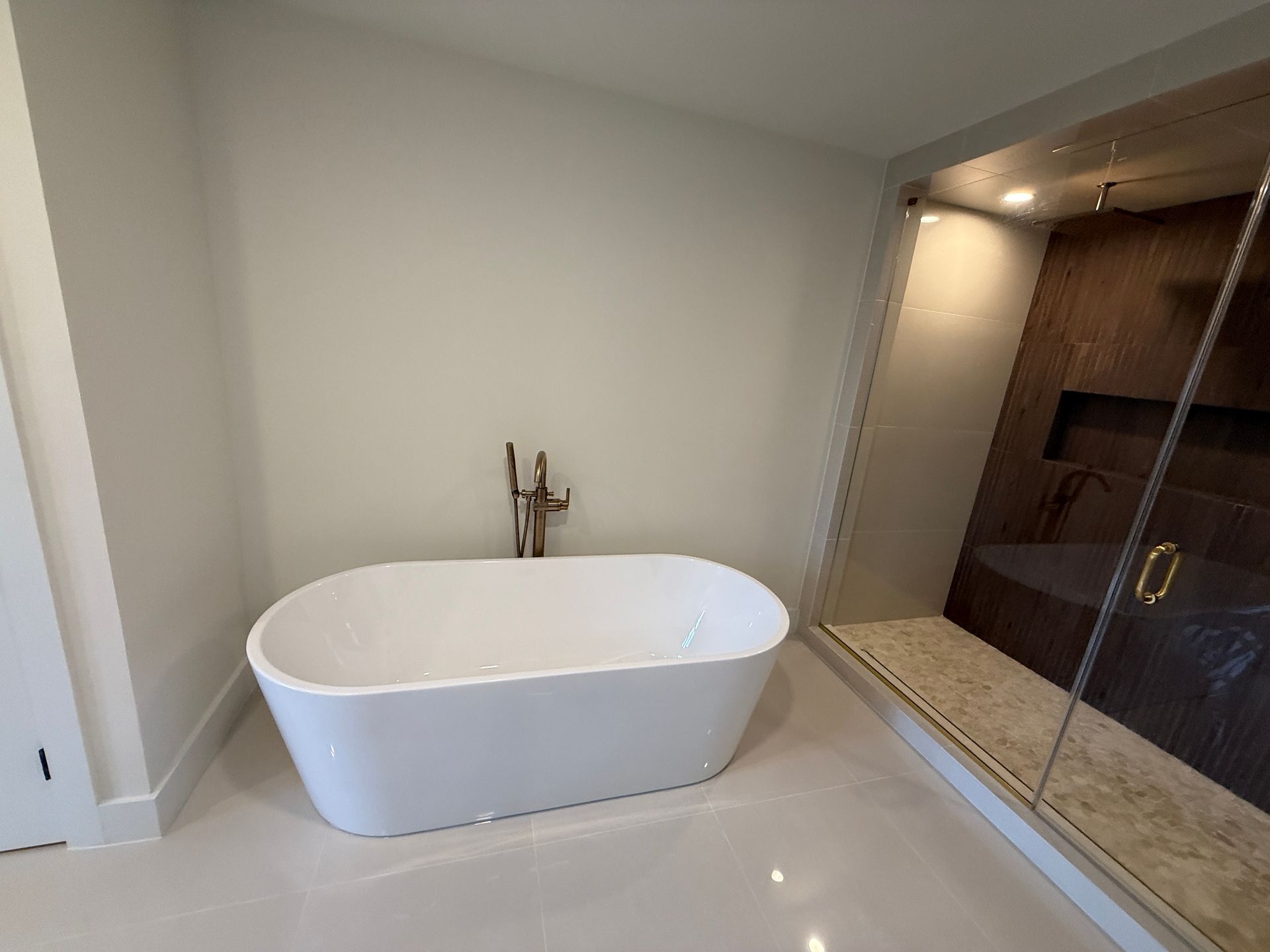 A freestanding white bathtub sits on a tiled floor next to a glass-enclosed shower with dark wood-paneled walls.