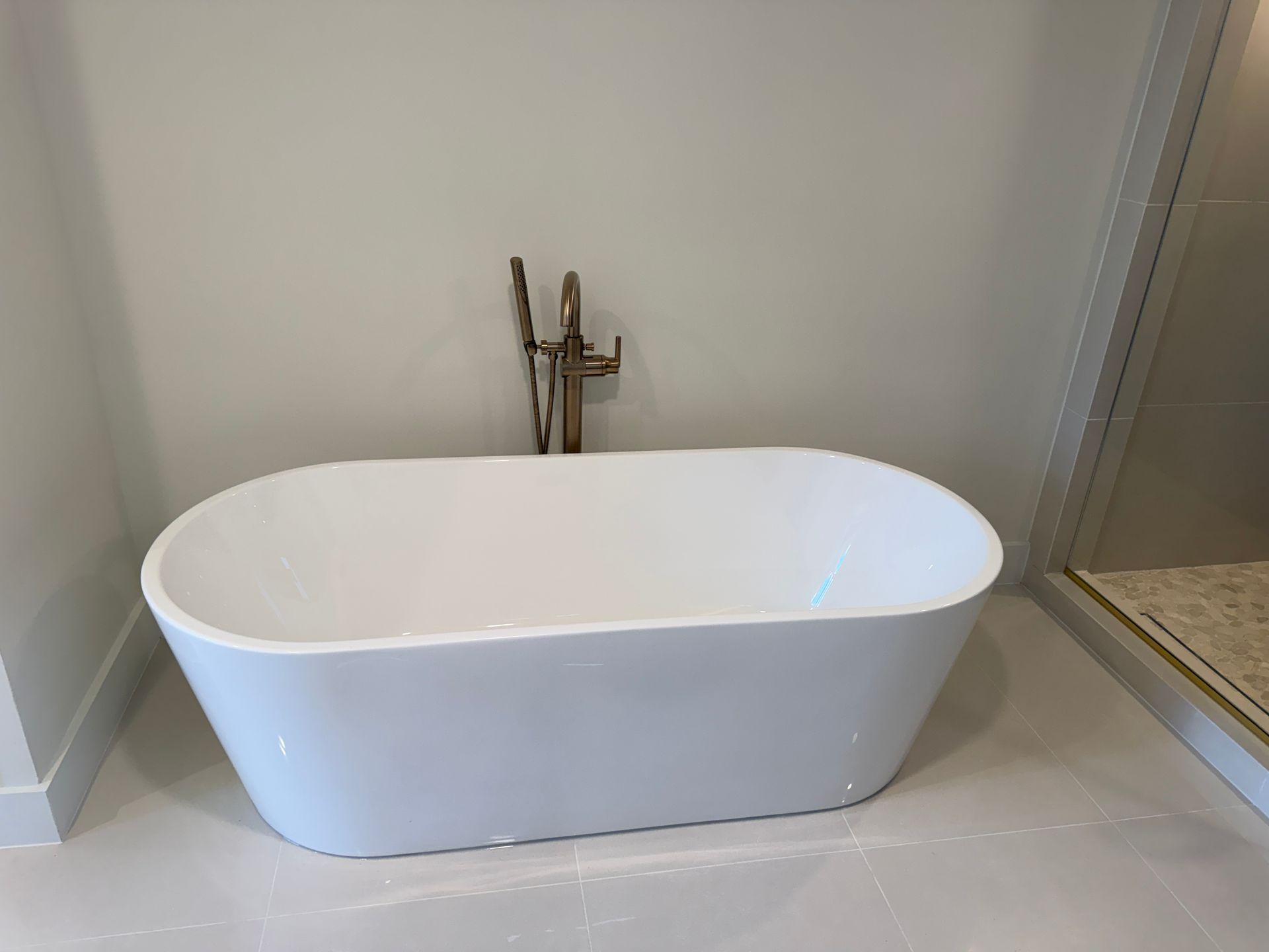 A white, oval freestanding bathtub sits in a modern bathroom with a gold floor-mounted faucet and tiled floors.