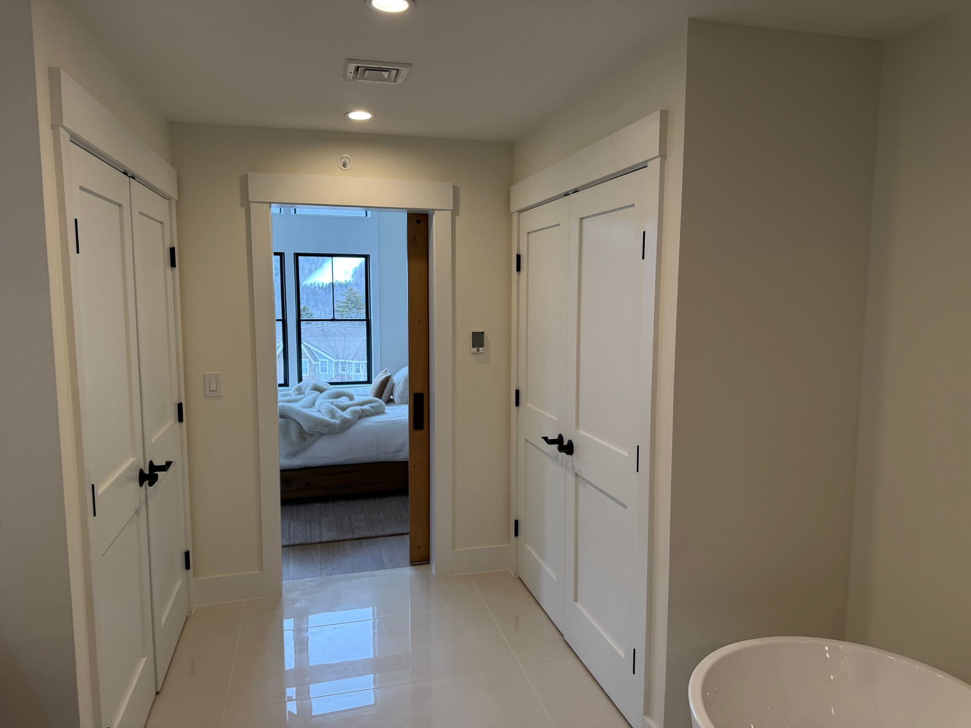 Hallway with white double doors on both sides, leading to a bedroom with a visible bed and a window.