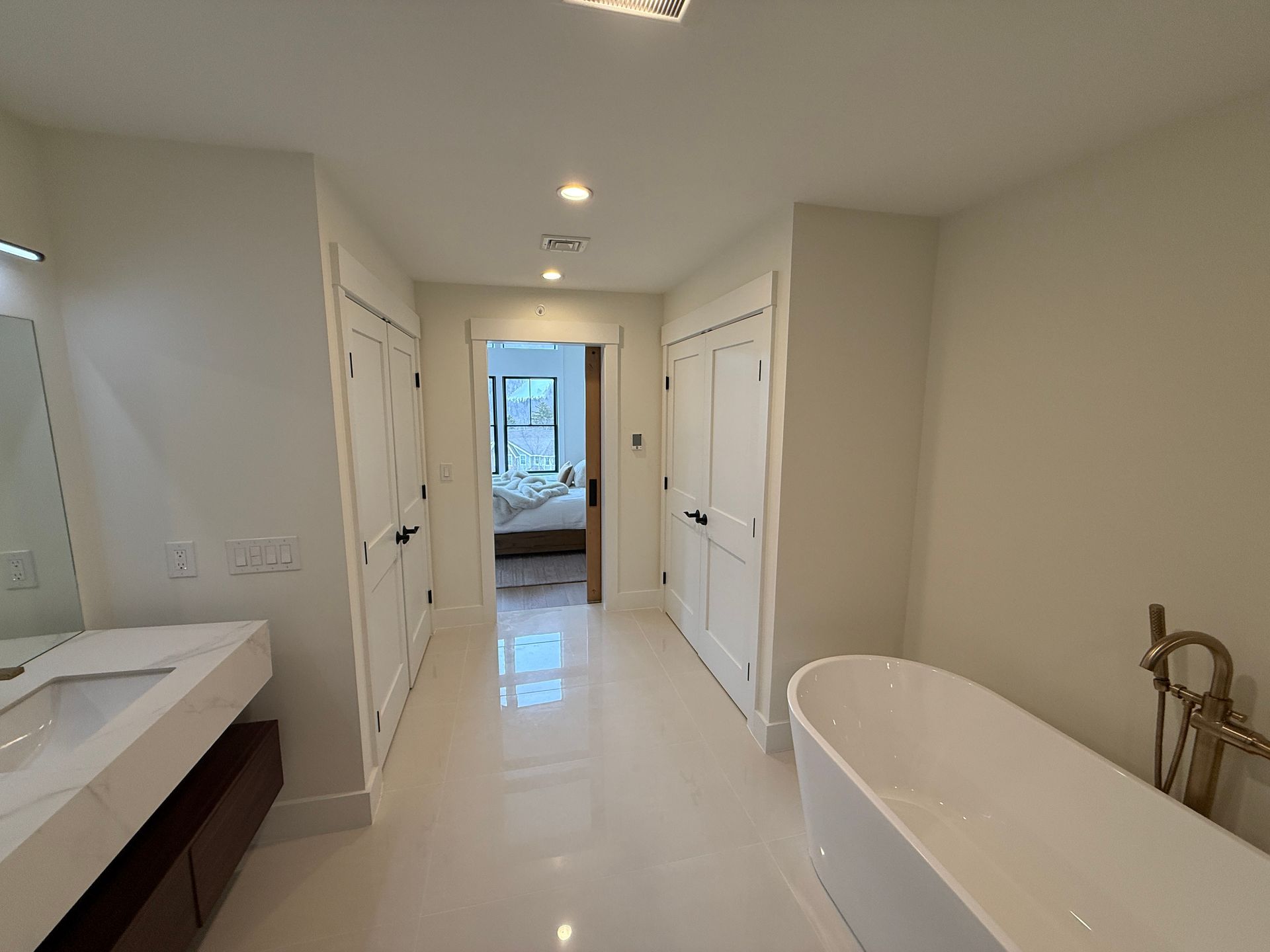 Modern bathroom with white walls, tiled floor, a freestanding tub, and a vanity, leading to an open bedroom doorway.