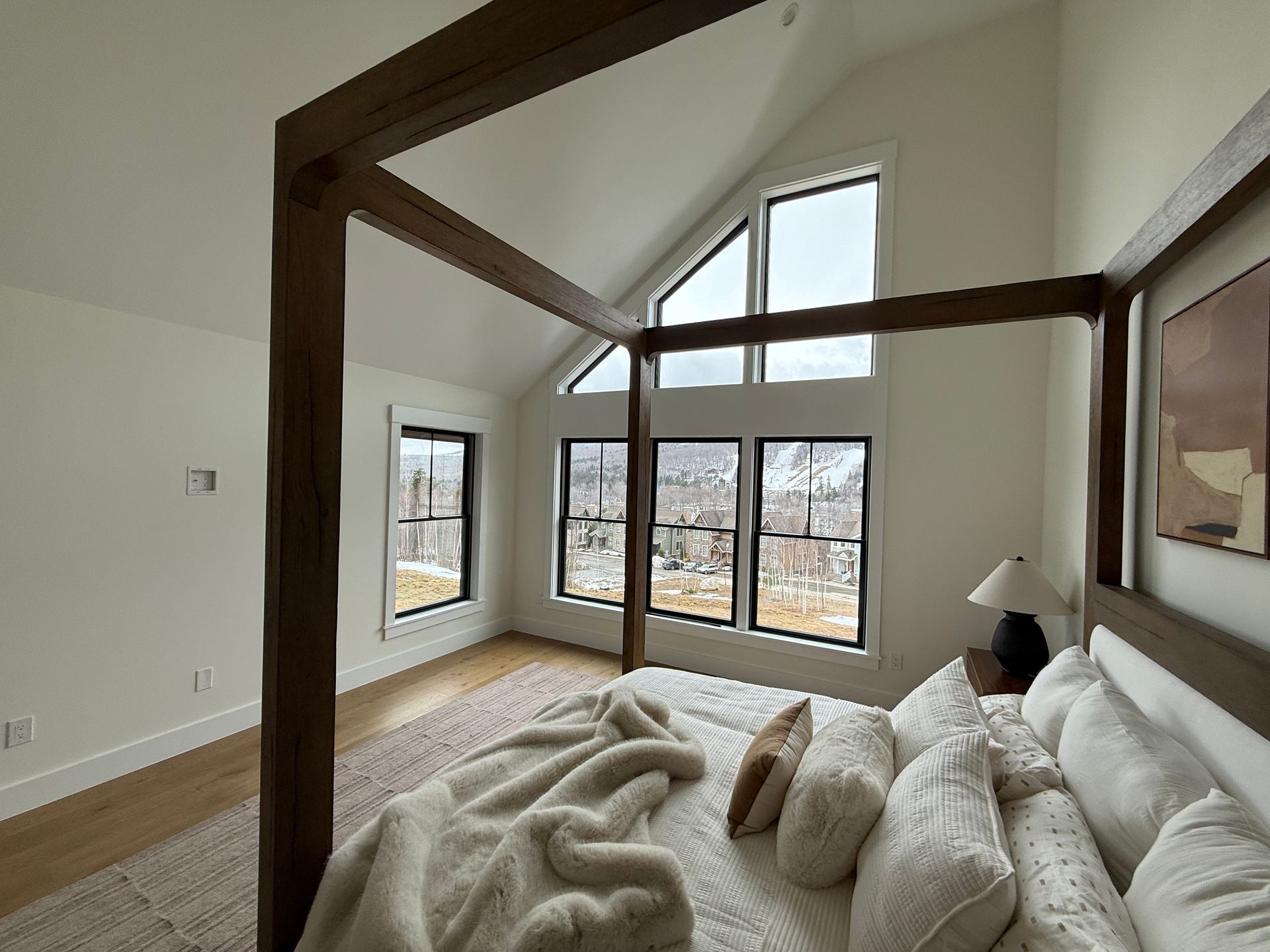 A light-filled bedroom with a wooden four-poster bed, white bedding, and large windows overlooking a snowy mountain view.