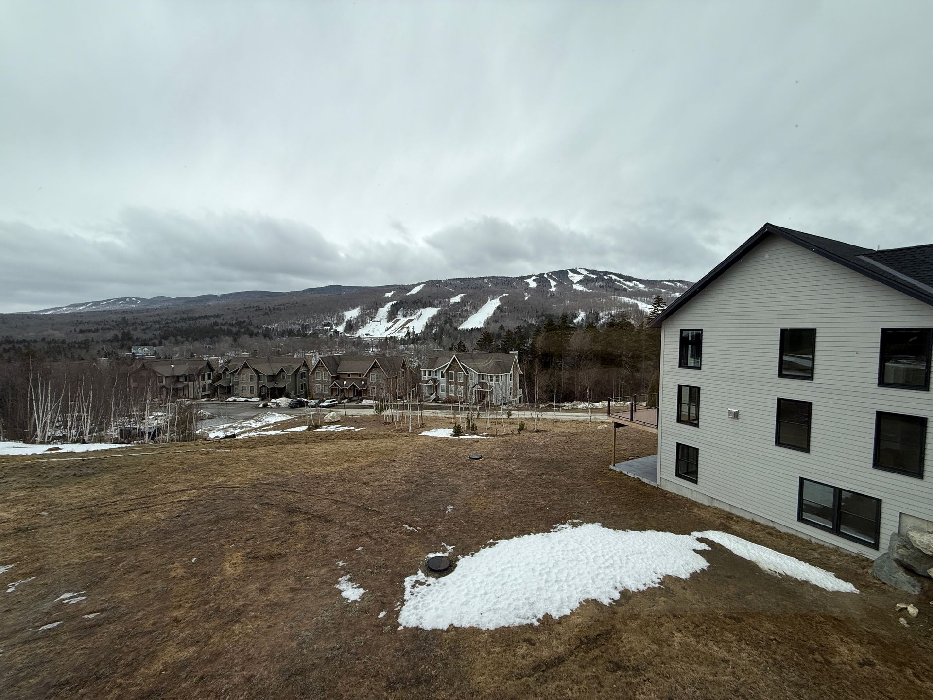 A light-colored multi-story building sits on a hillside near patches of snow, overlooking a mountain with ski slopes.