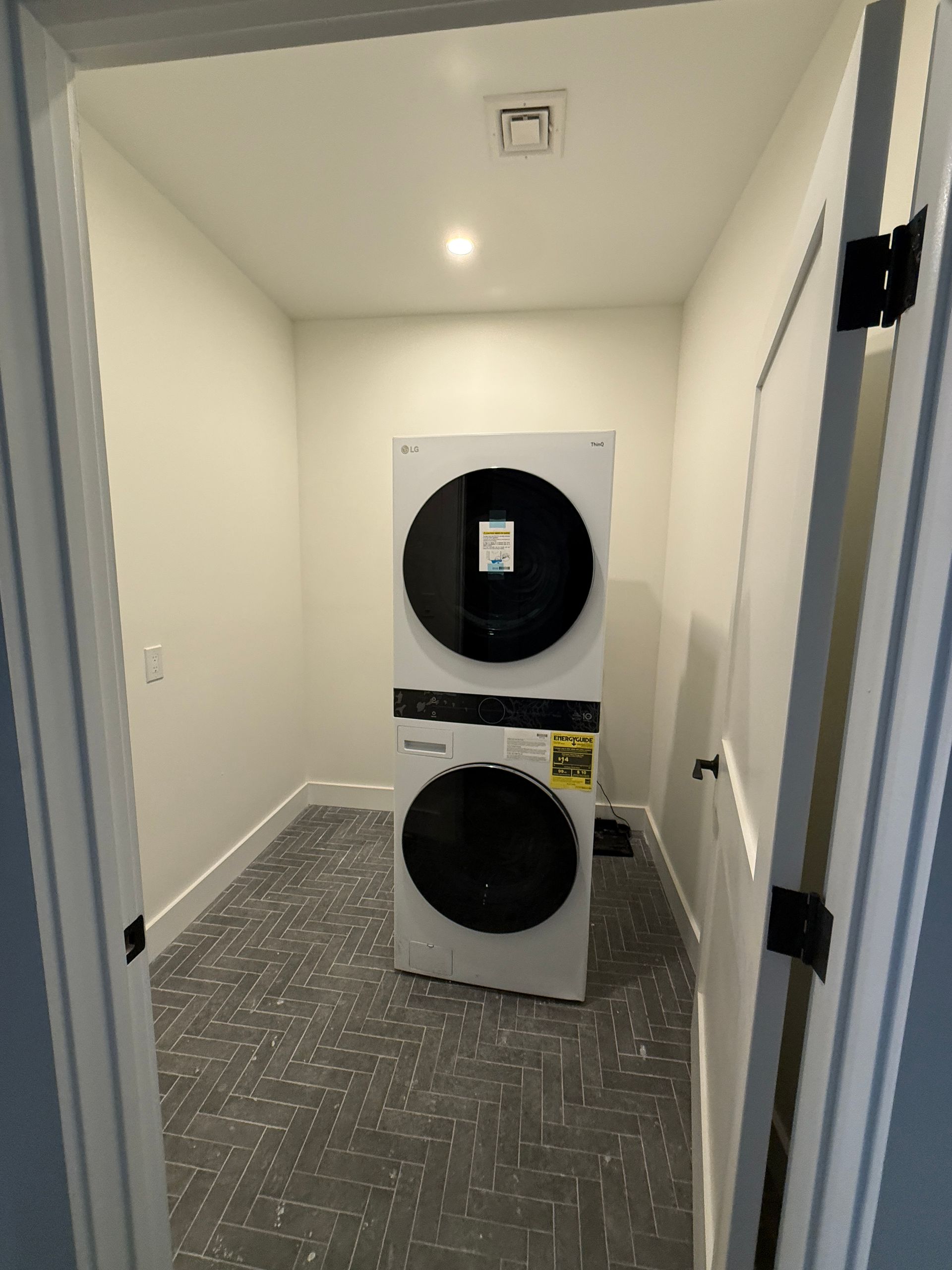 A white stacked washer and dryer unit stands in a small laundry room with gray patterned tile flooring and white walls.