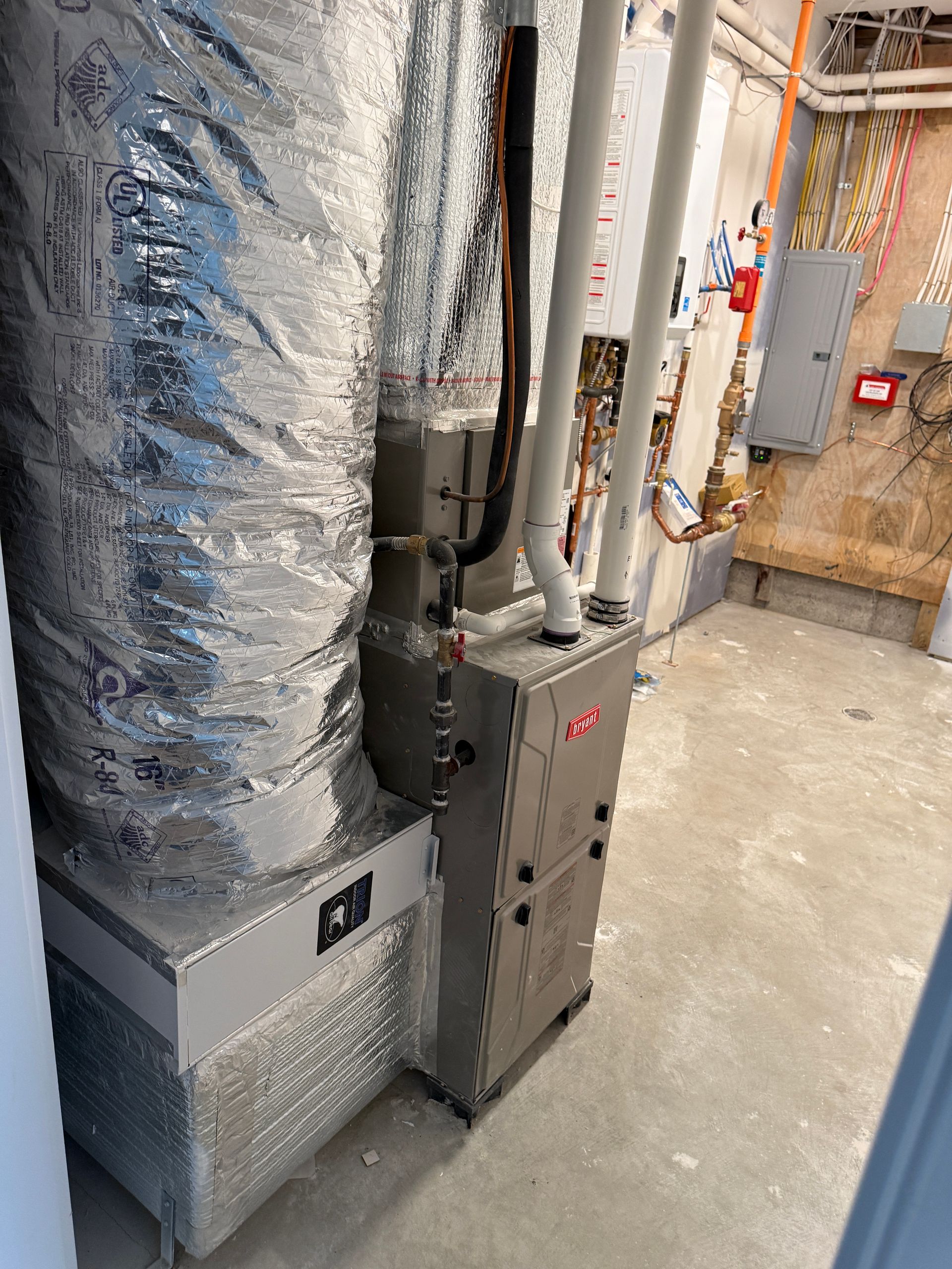 A residential HVAC furnace unit with silver ductwork in a basement mechanical room with concrete floors and wall panels.