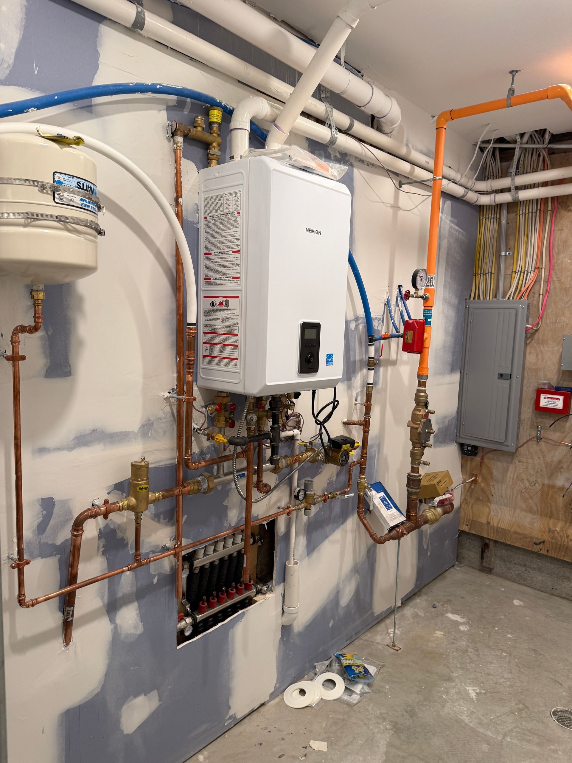 A wall-mounted boiler unit with copper plumbing, an expansion tank, and electrical panels in an unfinished basement room.