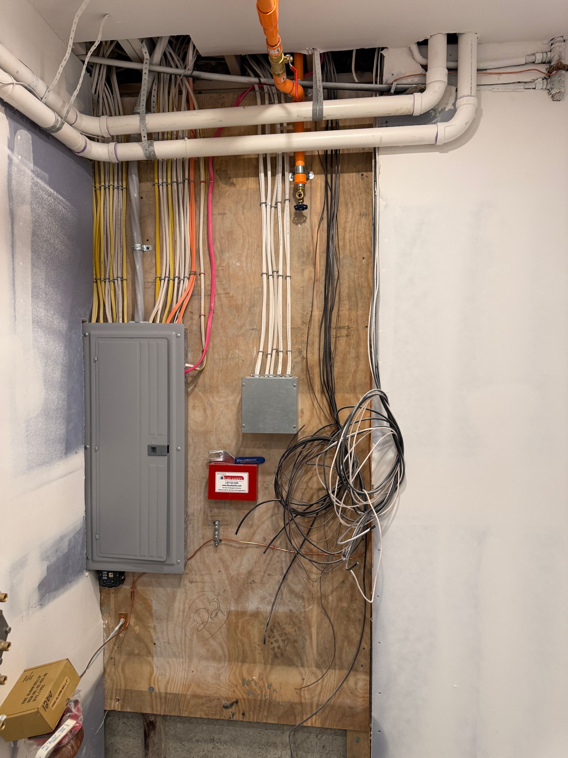 An electrical service panel and junction box mounted on a plywood wall with exposed wiring and overhead pipes.