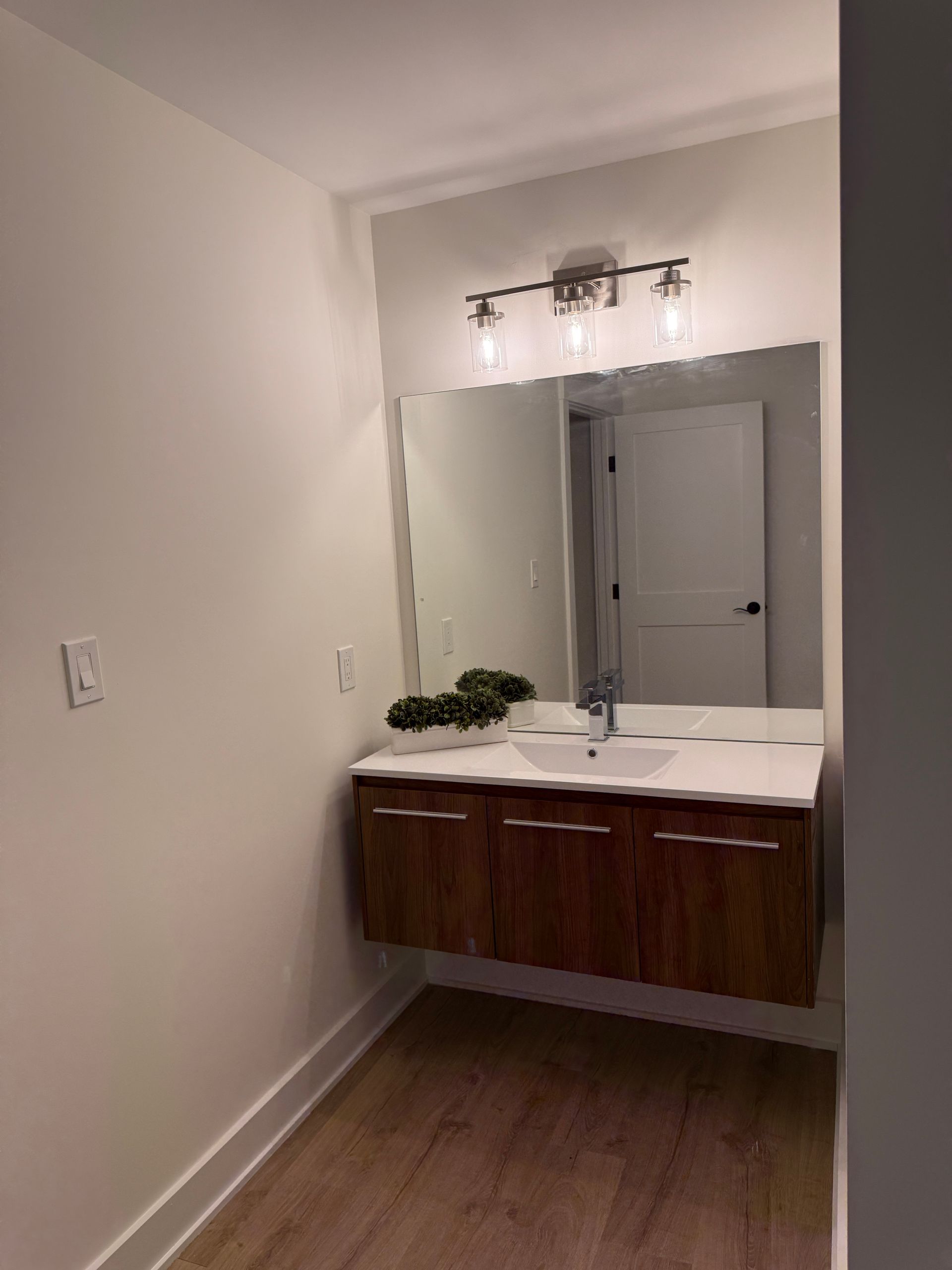 A modern floating vanity with a dark wood finish, large mirror, and three-bulb vanity light in a light-walled bathroom.