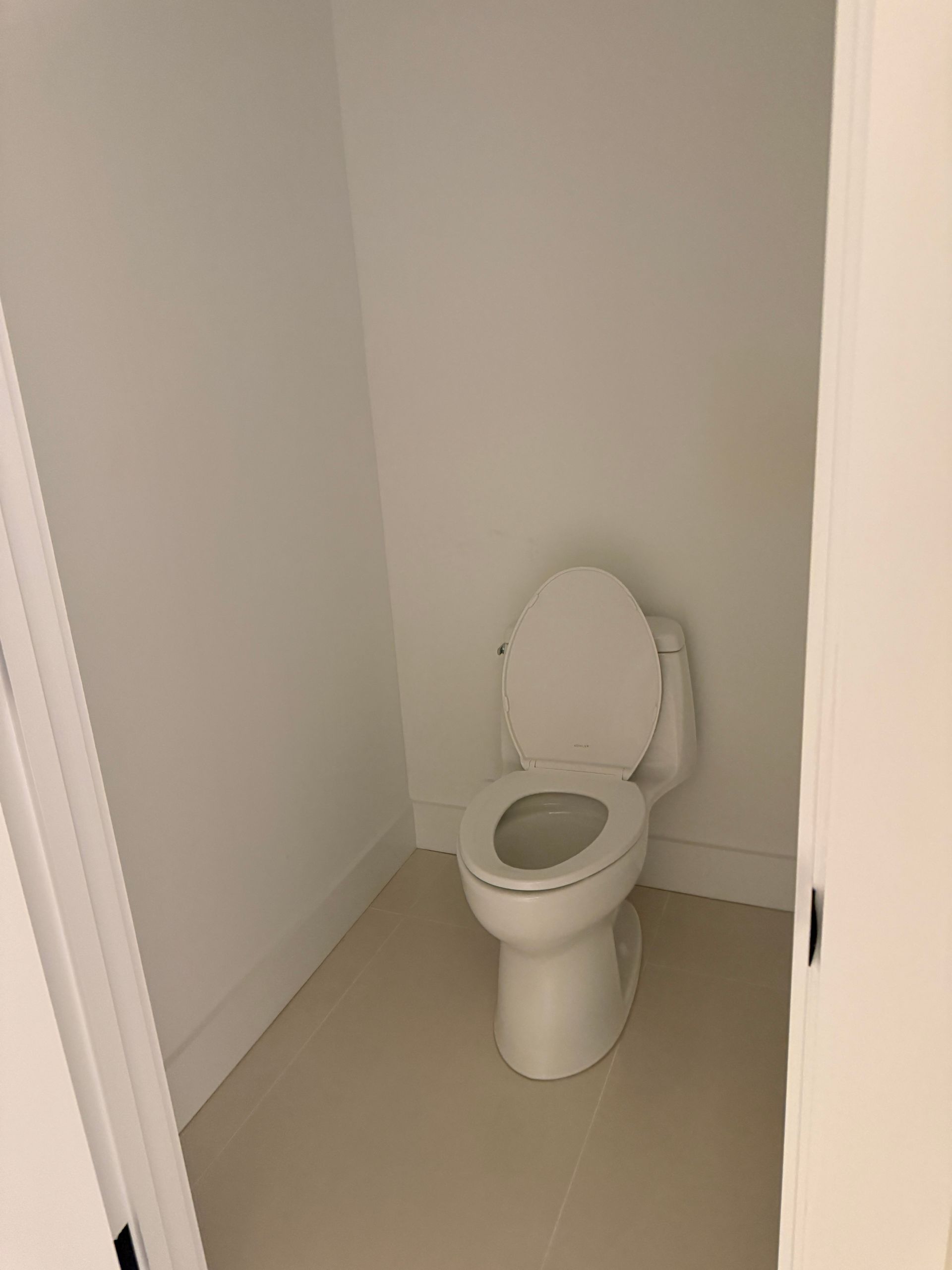A white toilet with the lid up inside a small, minimalist bathroom with white walls and light-colored floor tiles.