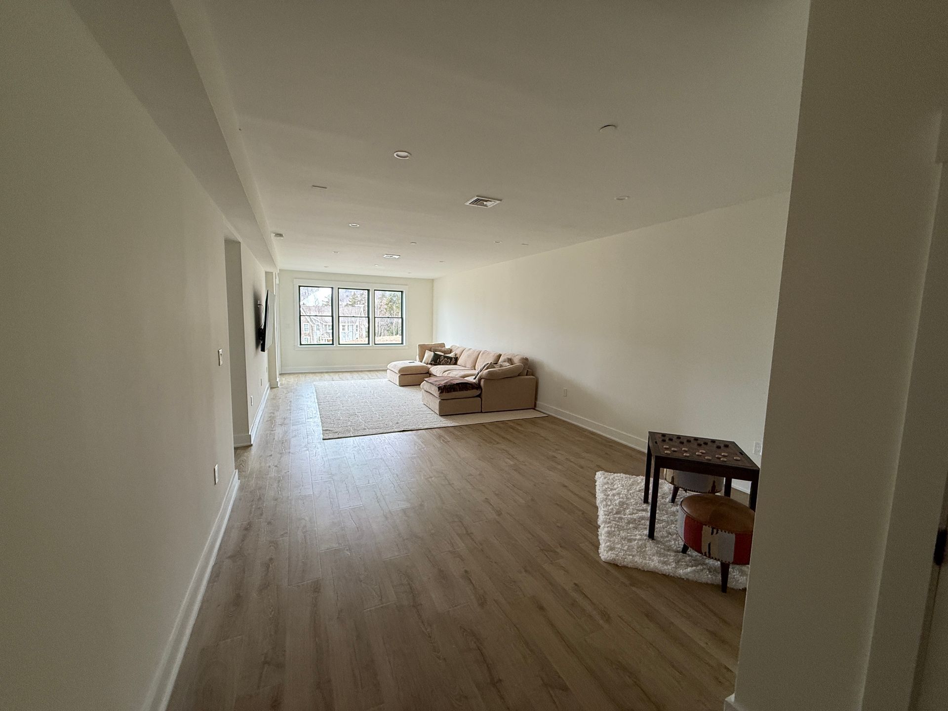 A wide view of a spacious living room with light wood floors, white walls, a beige sectional sofa, and a small table.