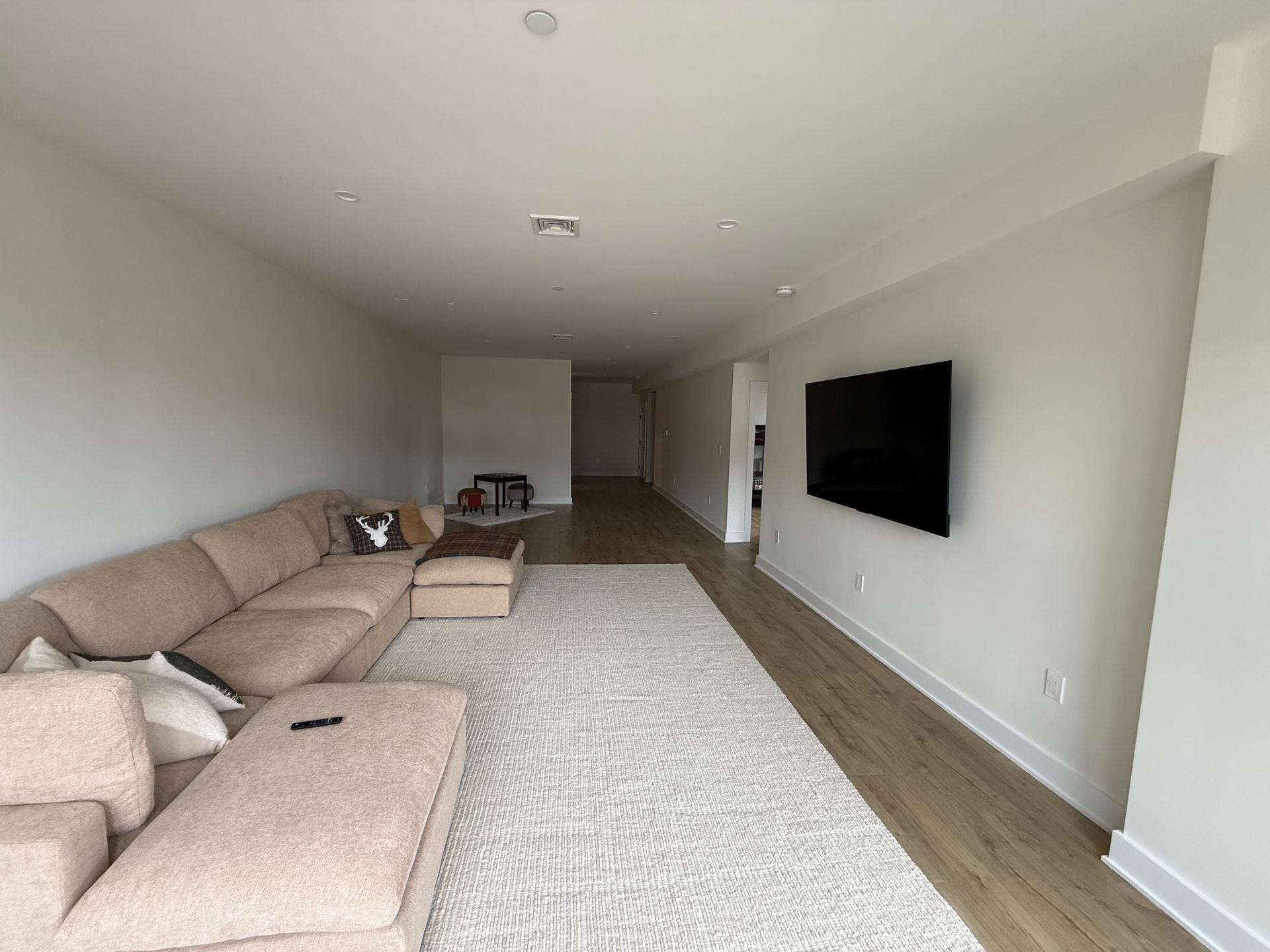 A long, minimalist living room with a large tan sectional sofa, light wood floors, a textured rug, and a wall-mounted TV.