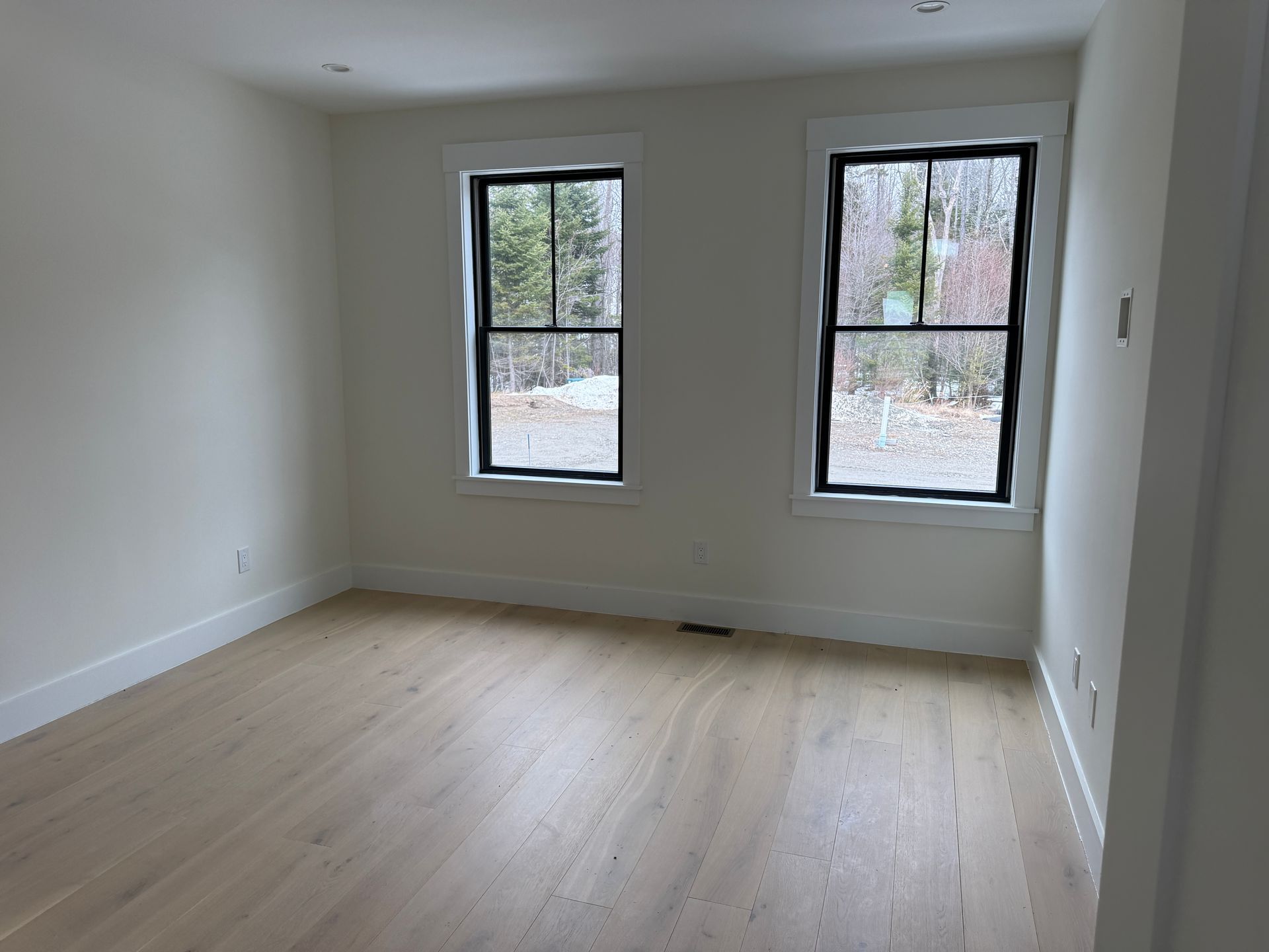 An empty room with light wood floors, off-white walls, and two windows looking out onto a wooded area.