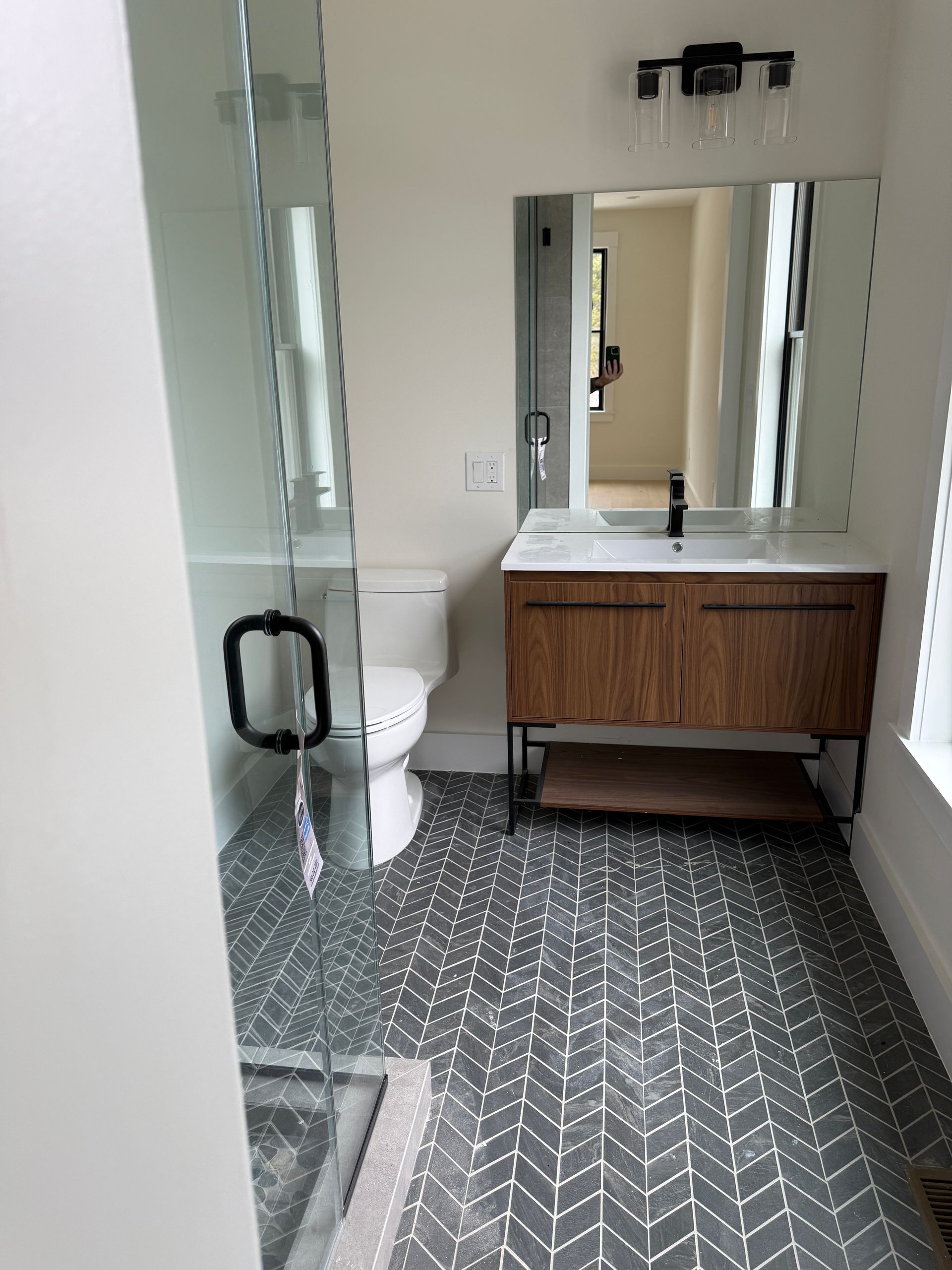 A modern bathroom with a glass-enclosed shower, wood vanity, white toilet, and gray-and-white chevron tiled floors.