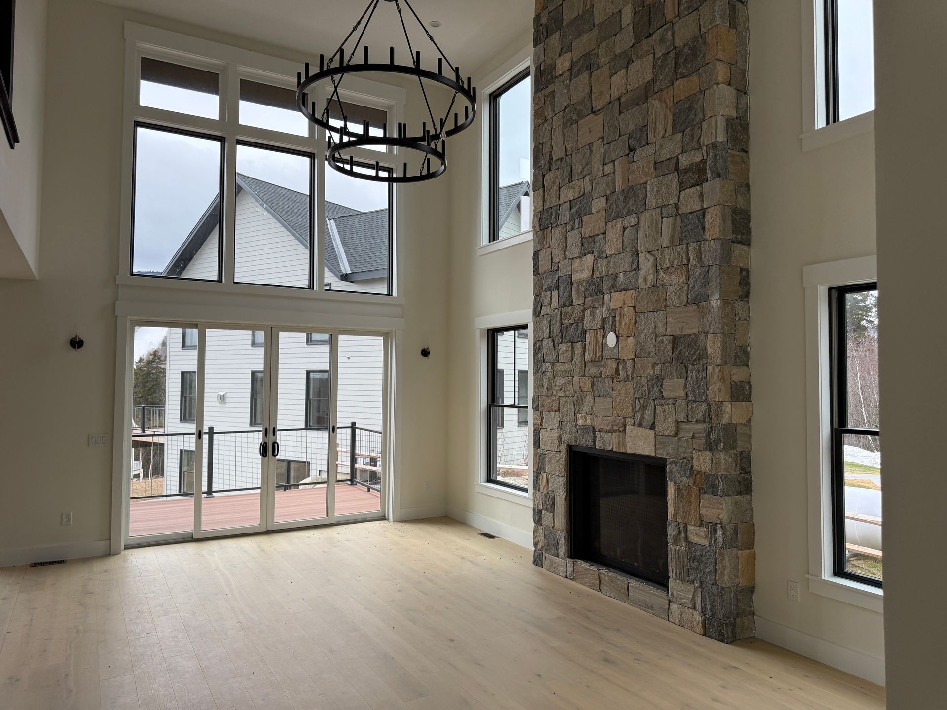 A living room with light wood floors, a tall stone fireplace, a large chandelier, and floor-to-ceiling windows.