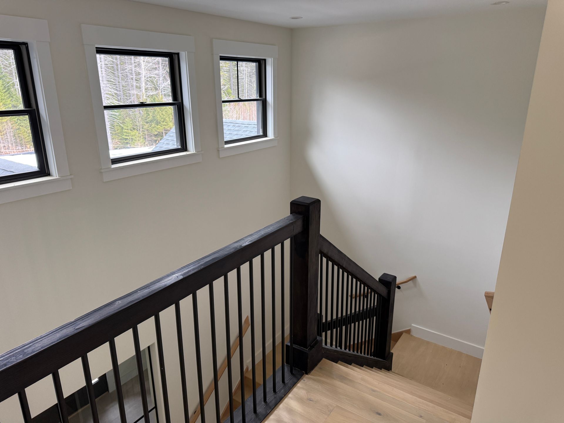 Wooden staircase with black railings descending along a white wall featuring three square windows.
