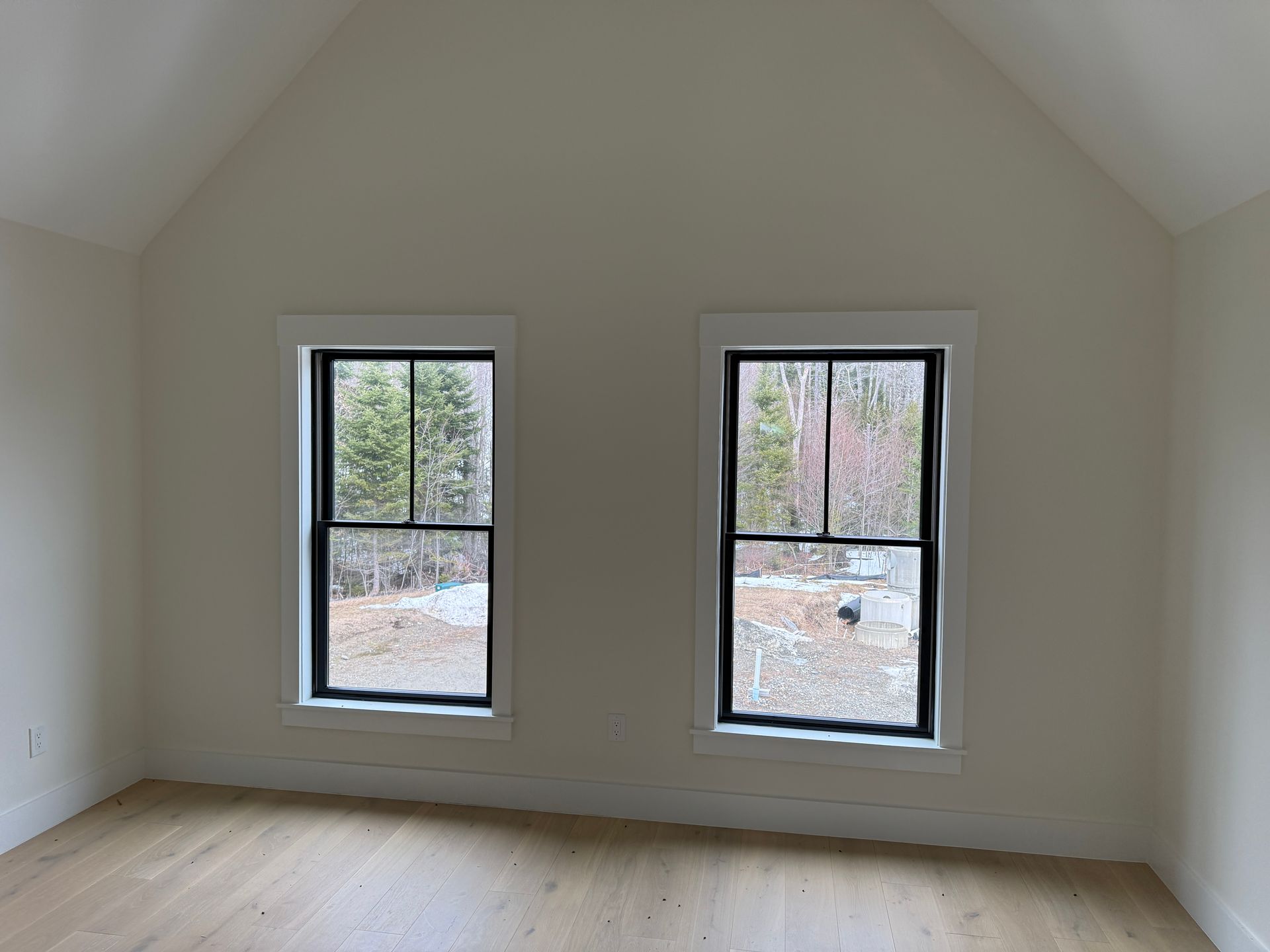 An empty room with light wood floors, beige walls, a vaulted ceiling, and two windows with black frames.