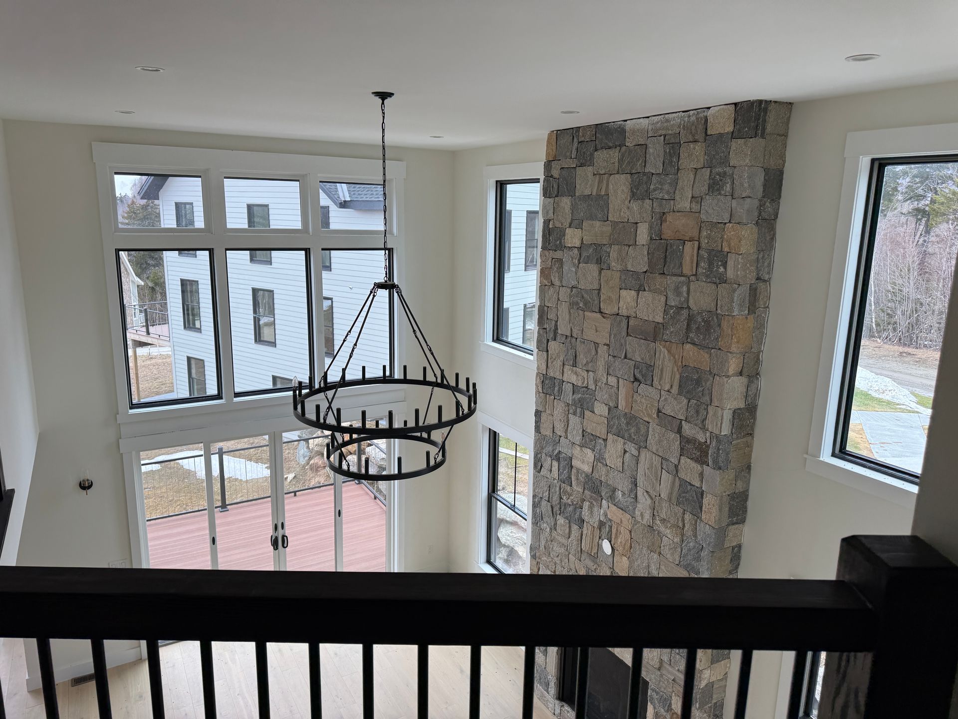 A high-ceilinged room with a tall stone fireplace, a tiered wagon-wheel chandelier, and large windows overlooking a yard.