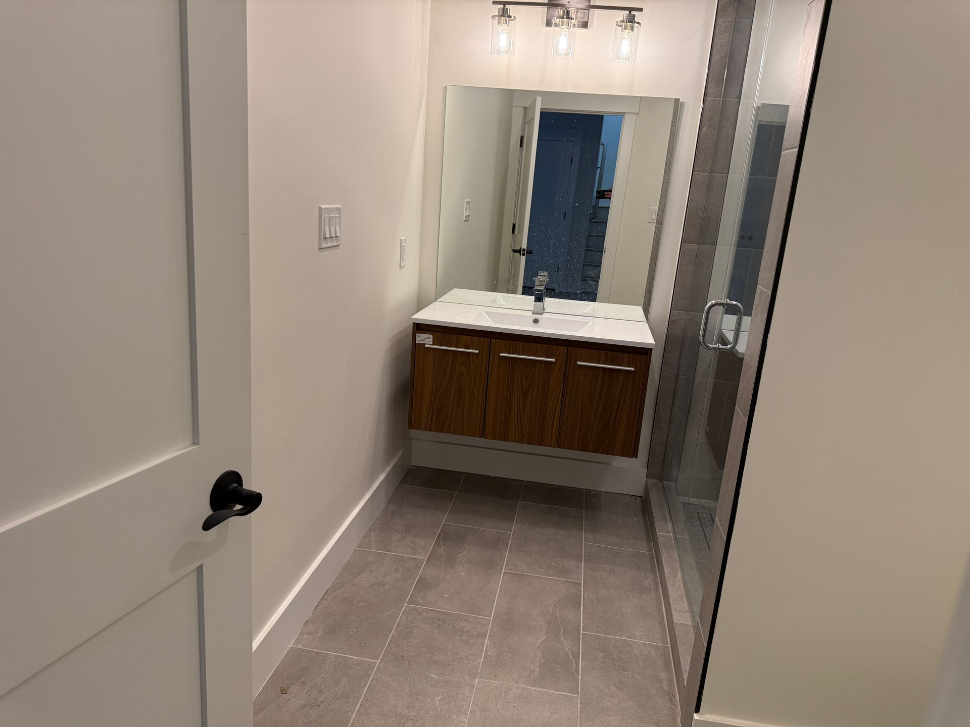 Modern bathroom with a floating wood vanity, white countertop, large mirror, glass-enclosed shower, and gray tile floor.
