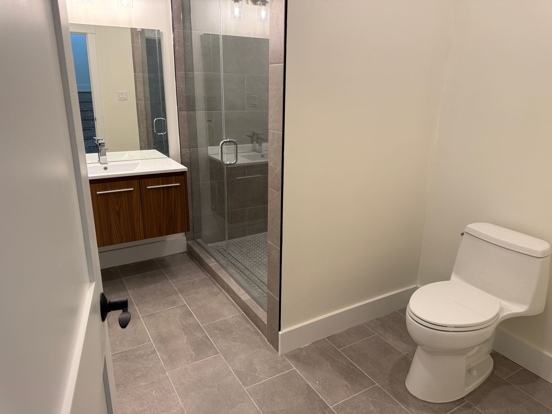 A modern bathroom with grey tiled floors, a wooden floating vanity, a glass-enclosed shower, and a white toilet.
