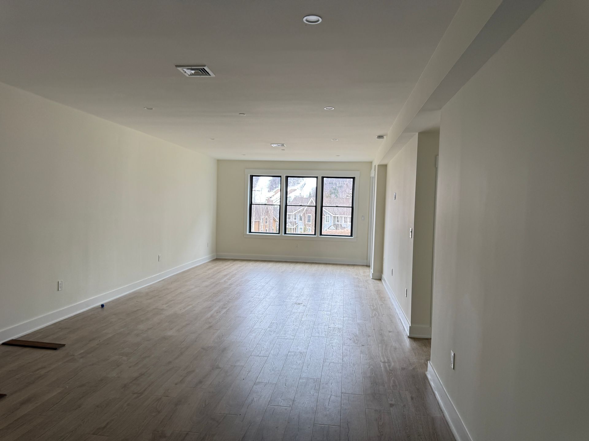 An empty room with light wood-tone flooring, white walls, recessed ceiling lights, and three windows at the far end.