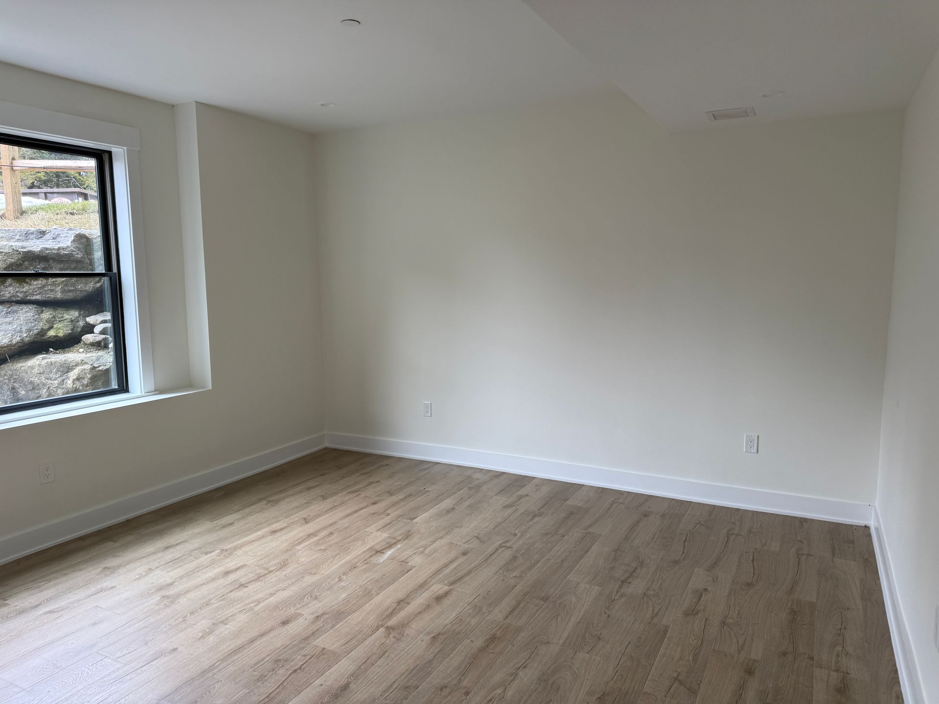 An empty room with light wood flooring, cream-colored walls, and a large window looking out onto a rocky landscape.